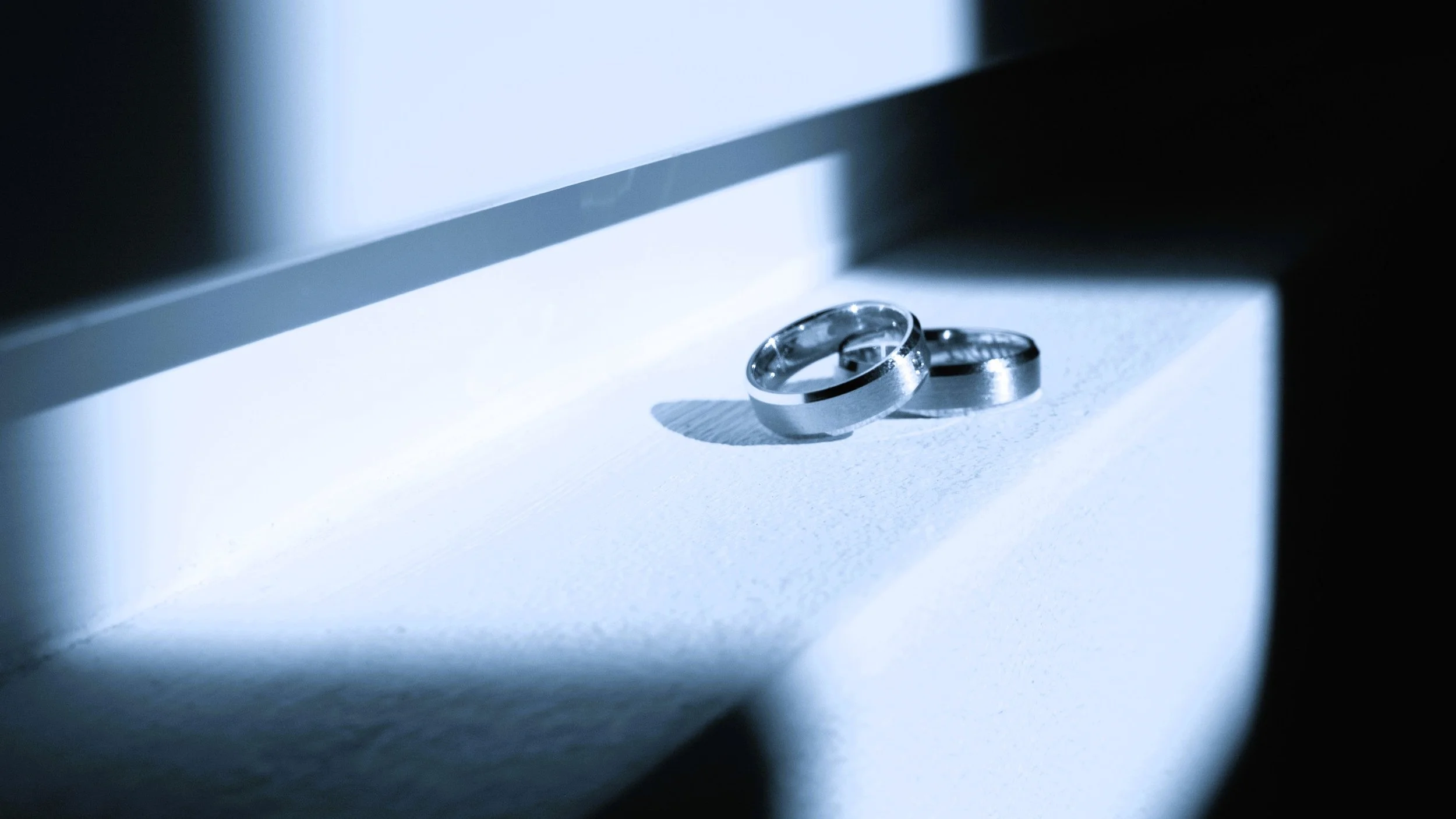 Two silver wedding rings placed on a white surface, with shadow and light playing across it, illuminated by a bright light source from above.