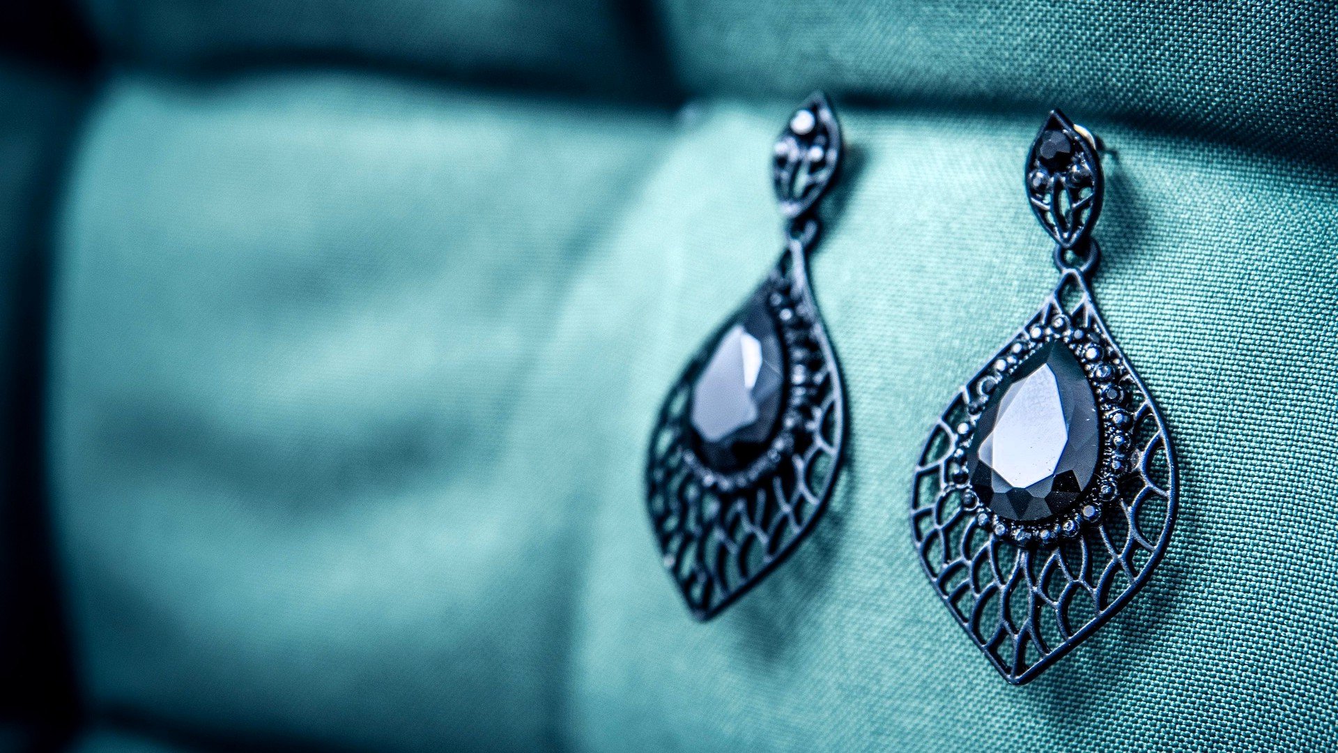 Close-up of black teardrop-shaped earrings with a lattice design and large central stones, resting on teal fabric.
