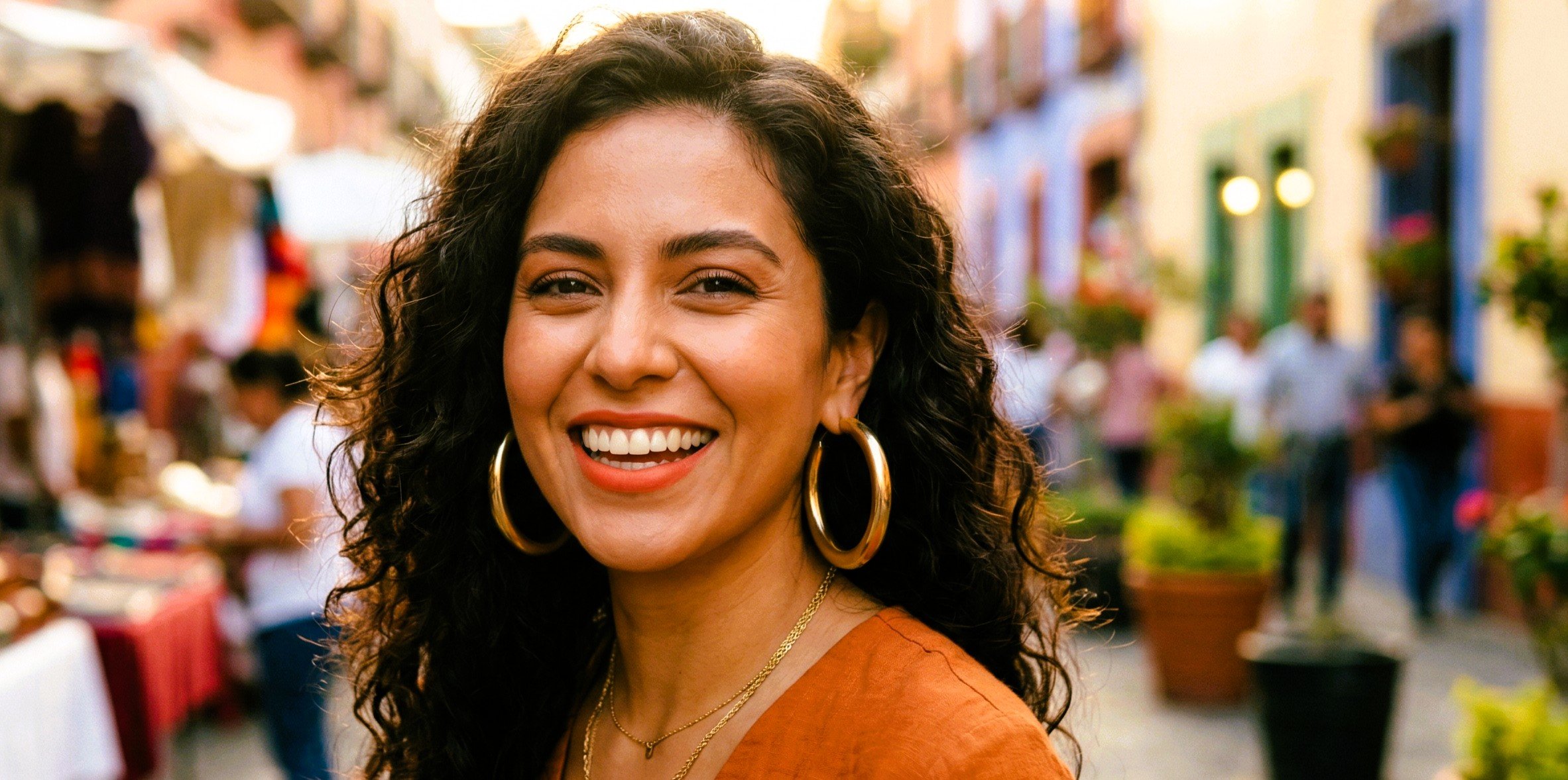 Smiling woman with curly black hair, gold hoop earrings, and a gold necklace at an outdoor market or street fair with colorful buildings and people in the background.