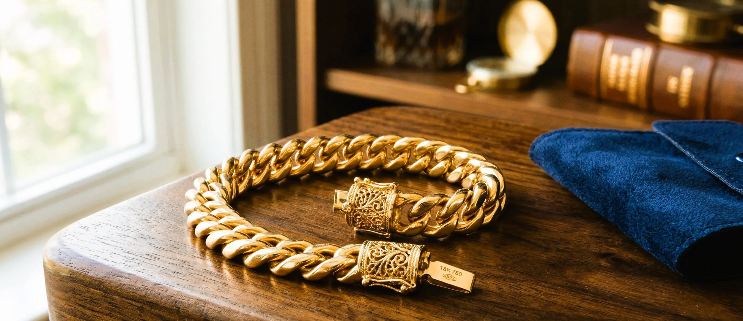 Close-up of a gold twisted chain necklace with ornate clasps on a wooden surface, next to a folded blue cloth and a book with gold accents in the background.