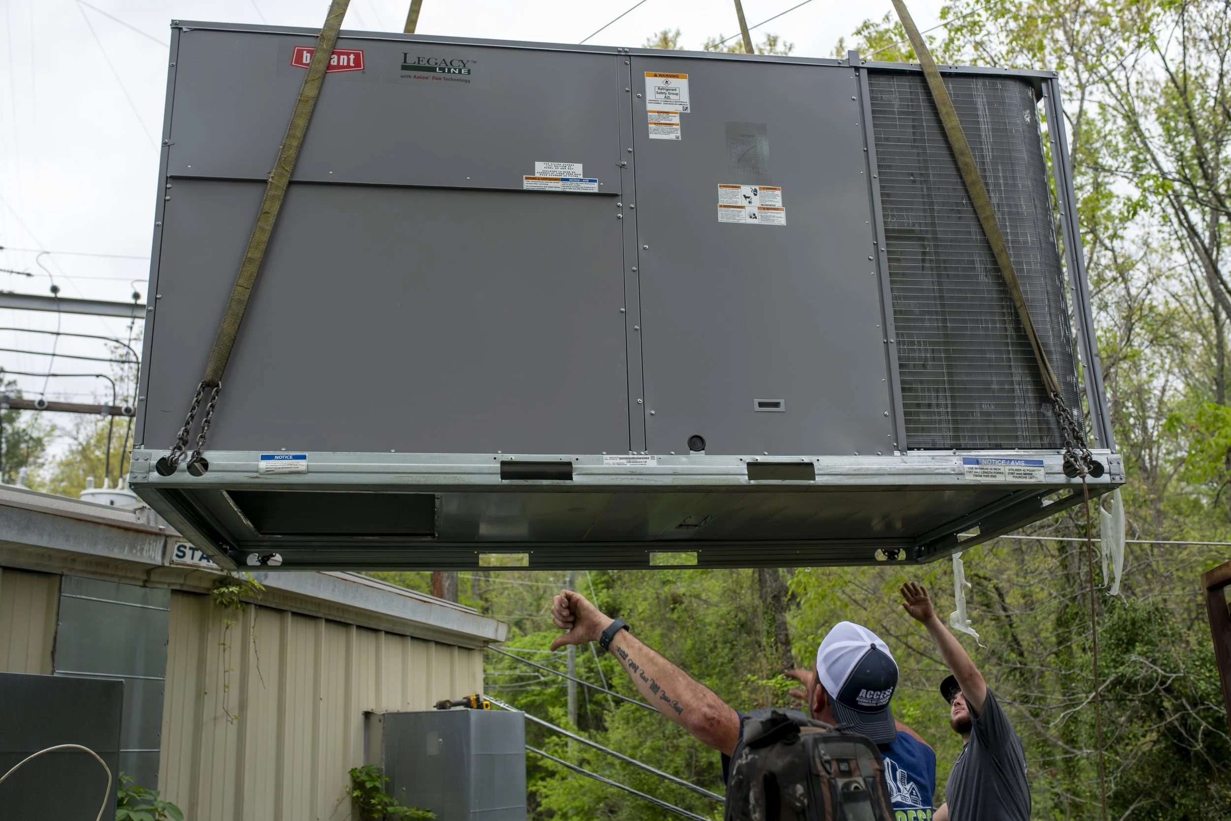 Two workers guide a large HVAC unit being hoisted by a crane outdoors, with trees and a building in the background.