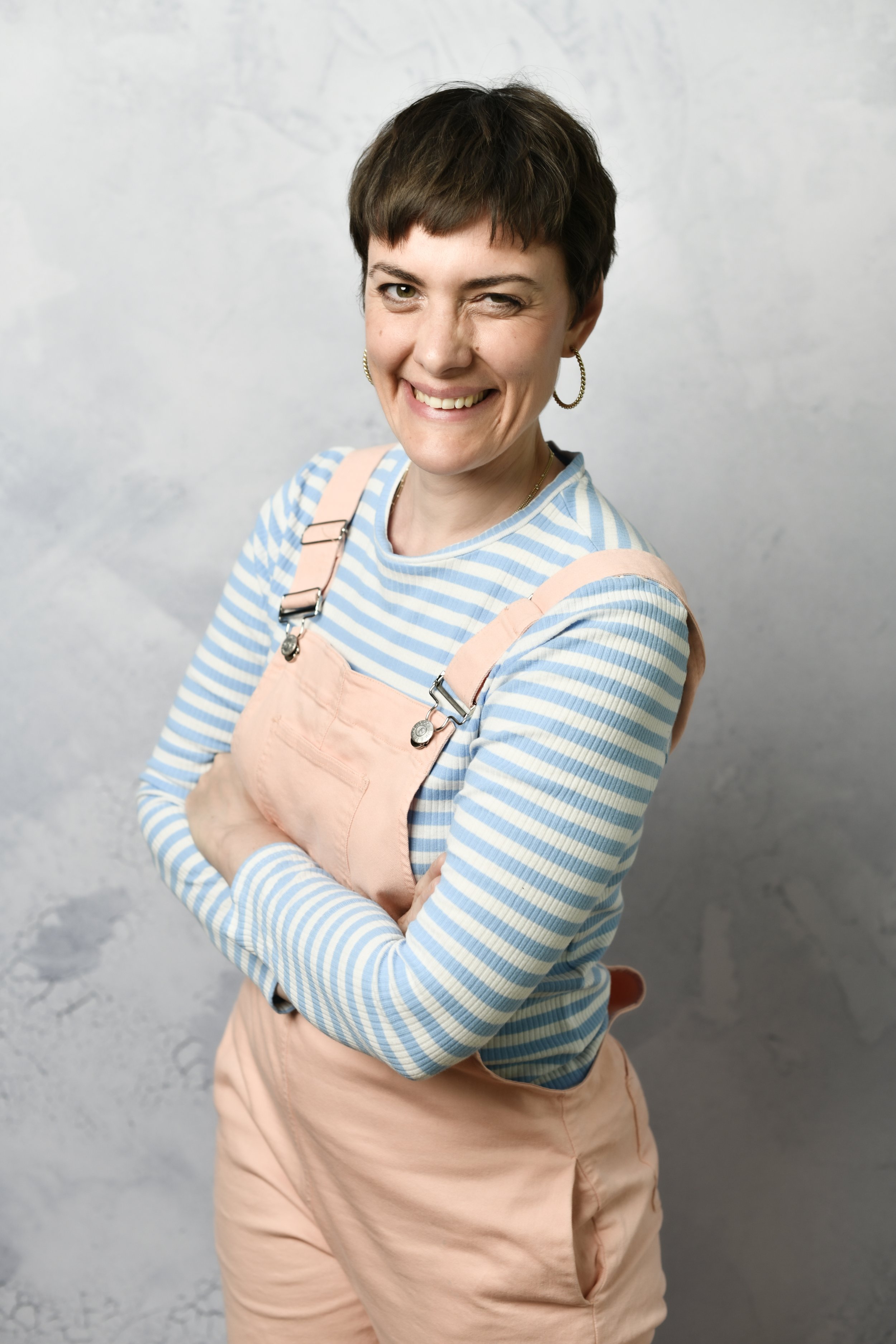 Junge Frau mit kurzen braunen Haaren, die einen Schulter-Wink-Wink zeigt. Sie trägt ein blau-weiß gestreiftes langärmeliges Hemd und rosa Latzhose, trägt ein Ohr-Hör.