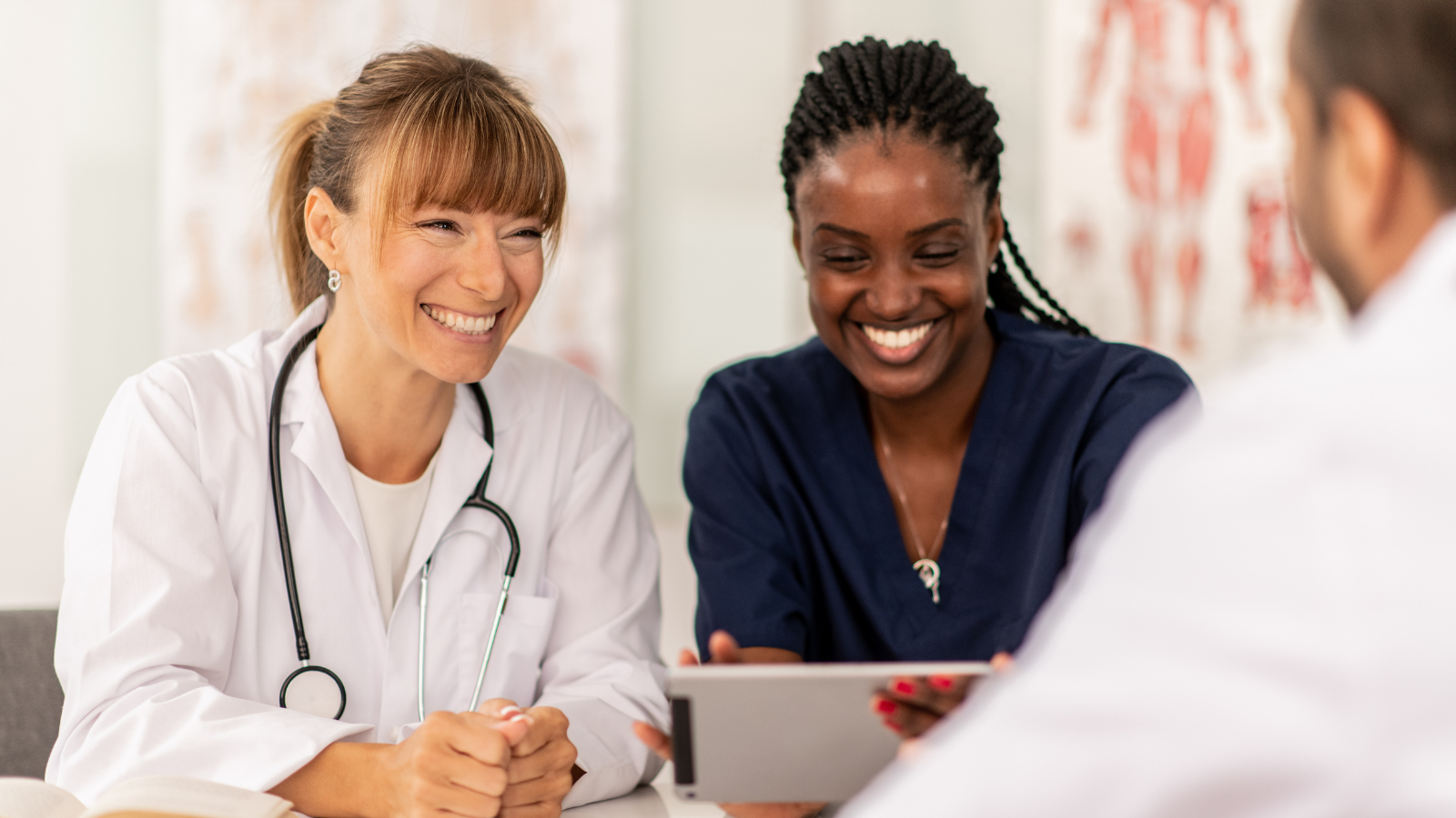 Two female healthcare professionals, one with a stethoscope, smiling and looking at a tablet, in discussion with a male colleague, in a medical setting with anatomical charts in the background.