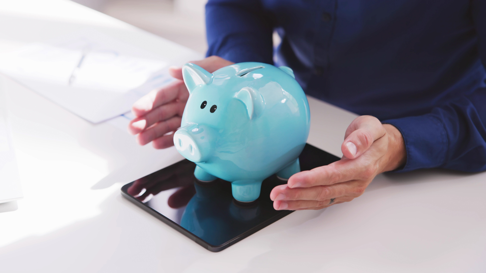 Person holding a blue piggy bank on a tablet, with papers and a laptop nearby.