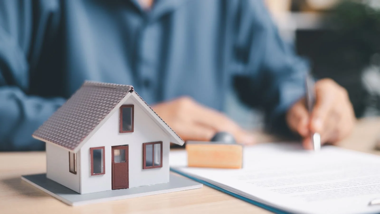 A miniature model of a house placed on a table with a person in the background writing or drawing on paper.