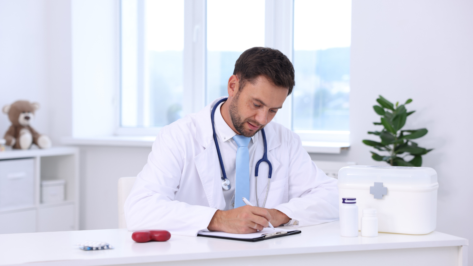 A male doctor in a white coat and a stethoscope around his neck sitting at a desk, writing in a notepad in a bright, modern medical office.