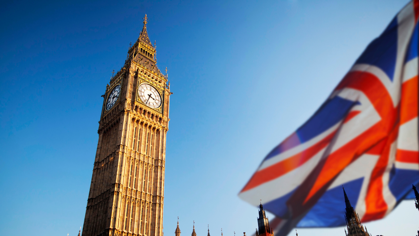 Big Ben clock tower with a partially visible British flag in the foreground, clear blue sky in the background.