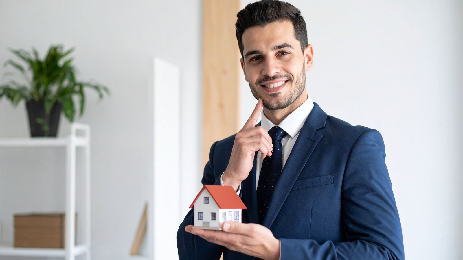 A man in a blue suit holding a small model house, smiling, in an indoor setting.