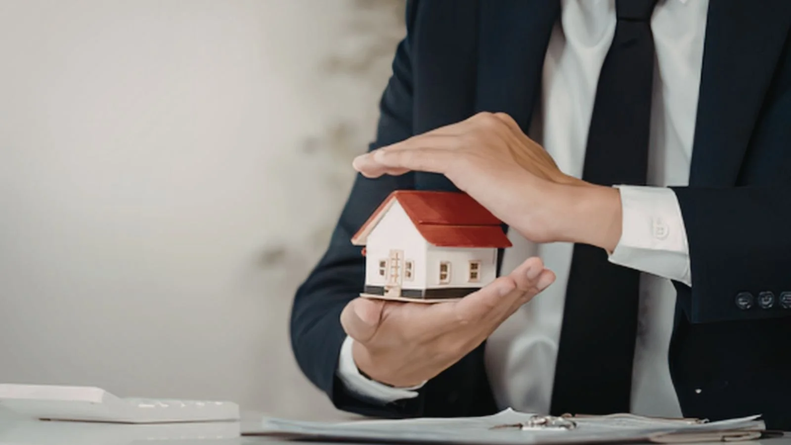 Person in a business suit holding a small model house with both hands.