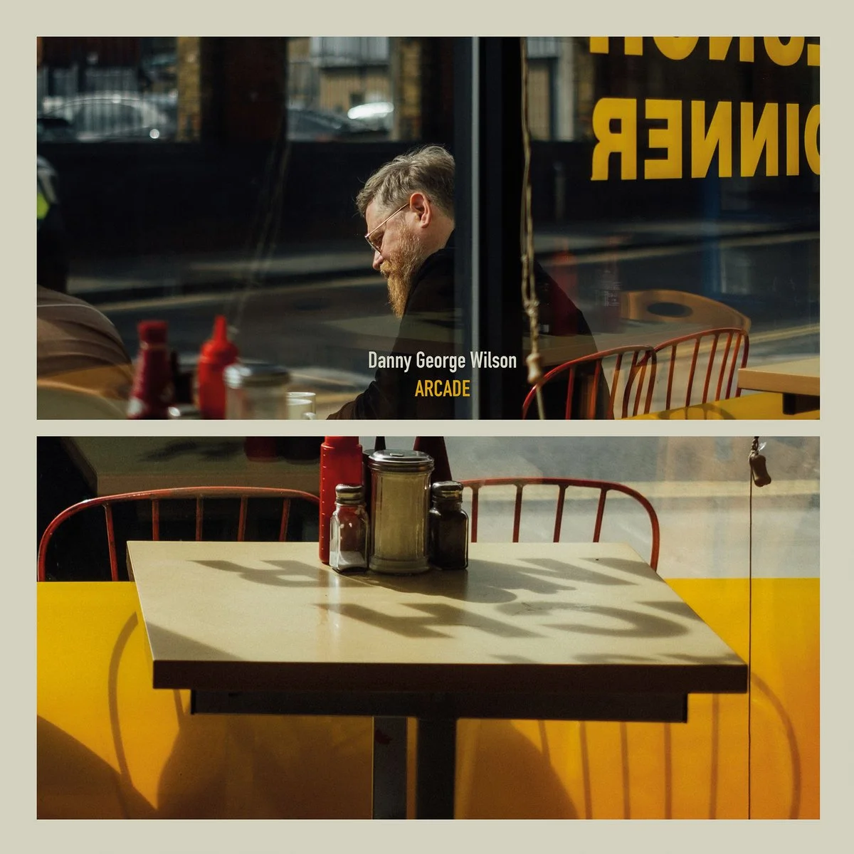 A man with glasses and a beard sitting inside a café, seen through a window, with condiment bottles on the table. The bottom part shows an empty table with yellow walls and red chairs outside the café