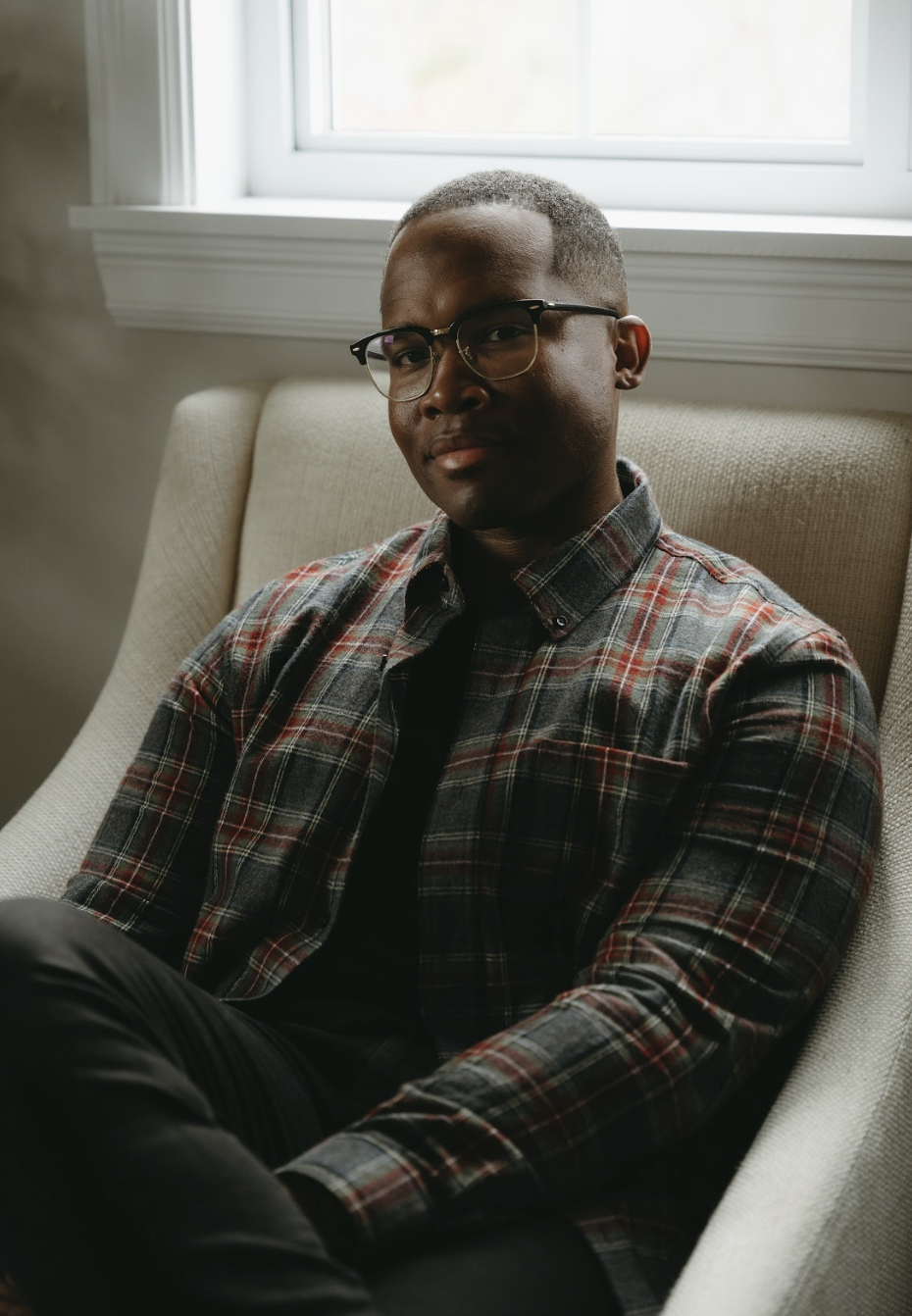 Marquis Murray wearing glasses and a plaid shirt sits on a beige armchair by a window.