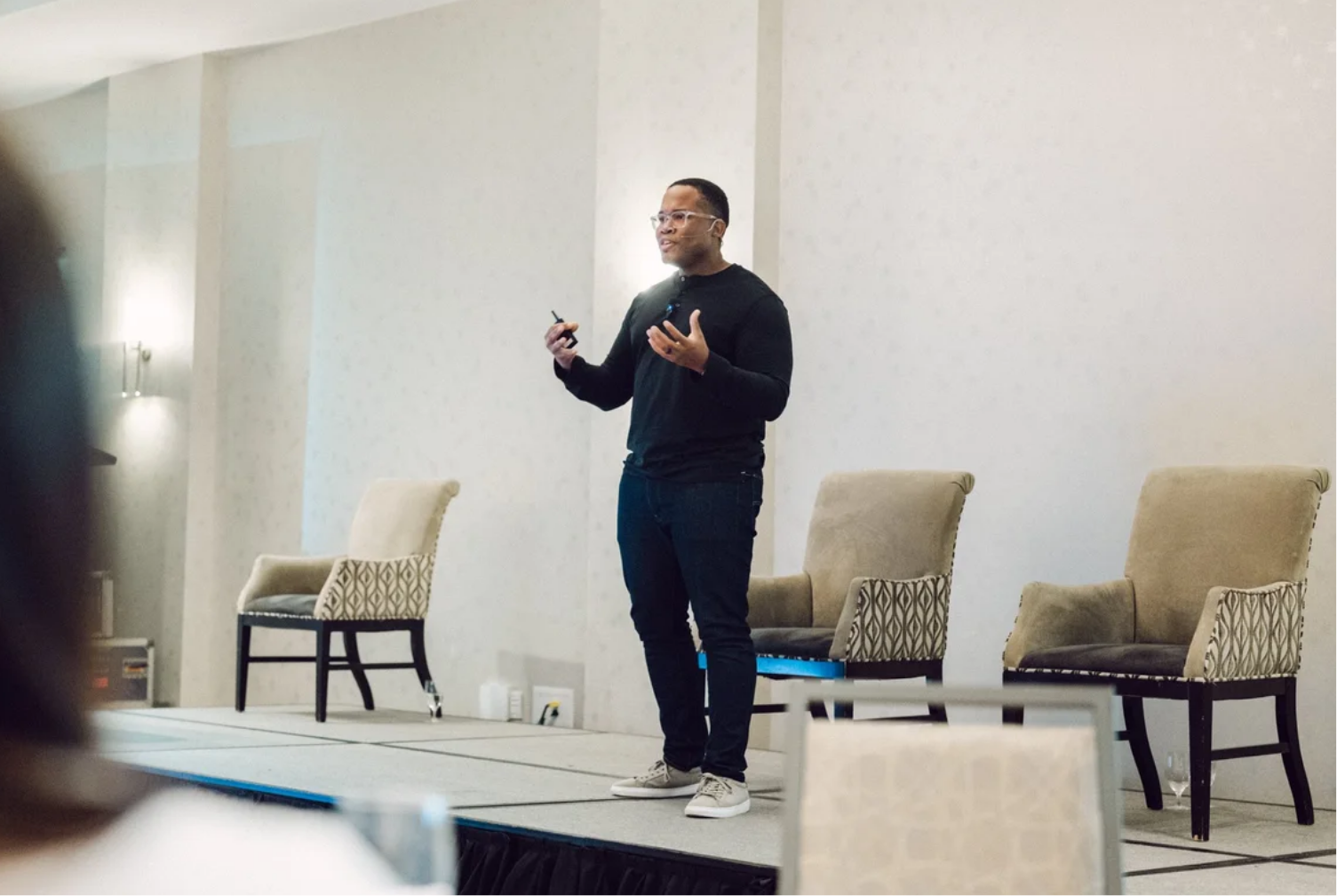 Marquis Murray dressed in black clothes and glasses standing on a stage, speaking to an audience. He holds a microphone and a clicker, with two beige chairs behind him. The room has a simple decor with soft lighting.