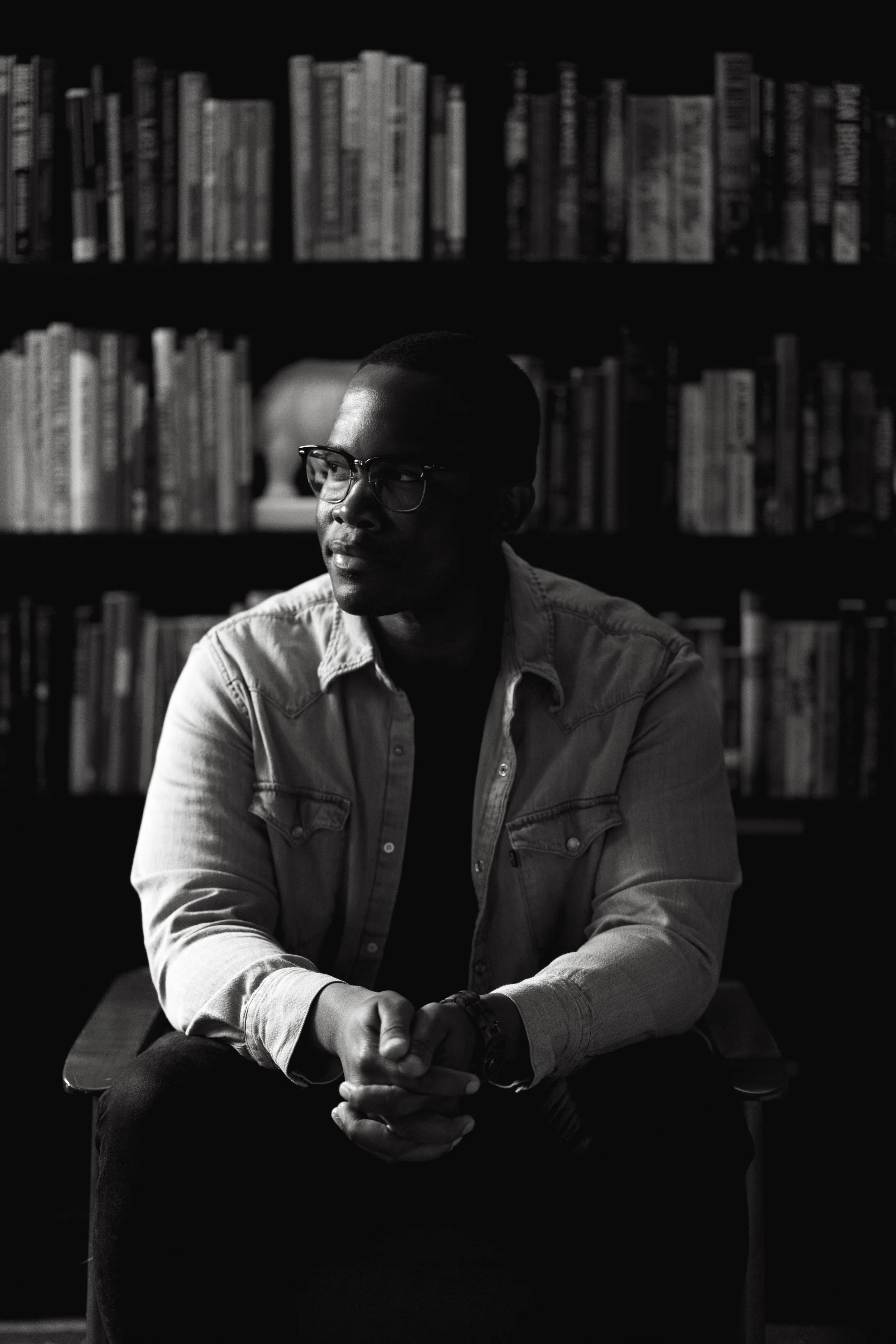 Marquis Murray with glasses, and short hair sitting at a wooden table in front of a background of bookshelves filled with books, captured in black and white.