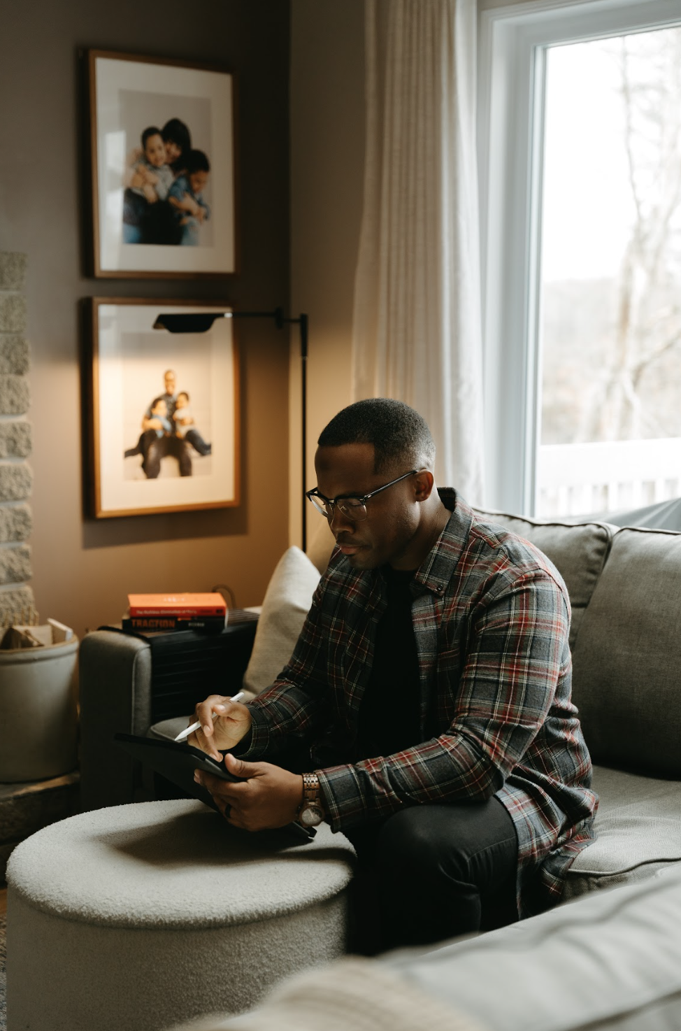 Marquis Murray sitting on a beige ottoman in a living room, using a tablet with a stylus. The room has a grey couch, a window with cream curtains, and framed photographs of his children on the wall.