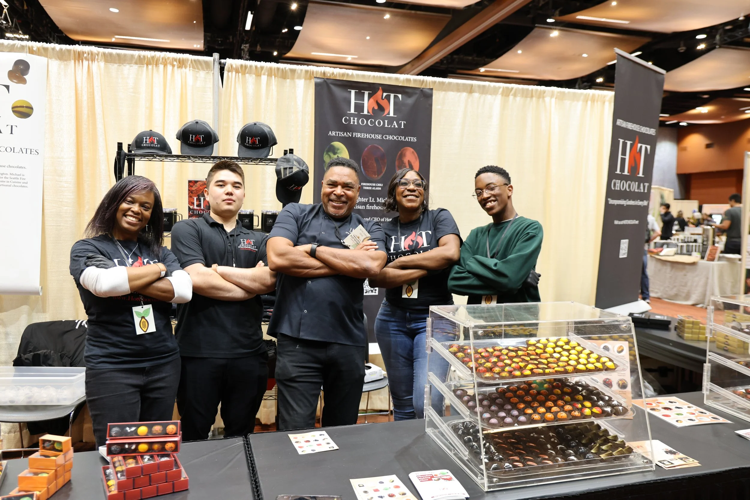 Group of five smiling people standing behind a display of assorted chocolates at a booth, with banners and hats promoting 'Hot Chocolat' artisan firehouse chocolates in the background.