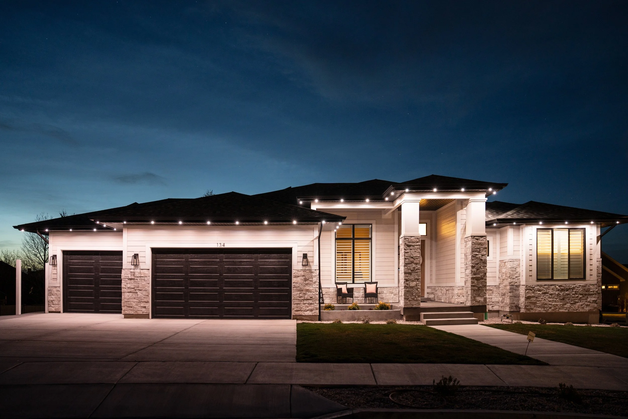 A modern single-story house illuminated at night, featuring a three-car garage, stone accents, and a covered porch with seating, under a dark sky.