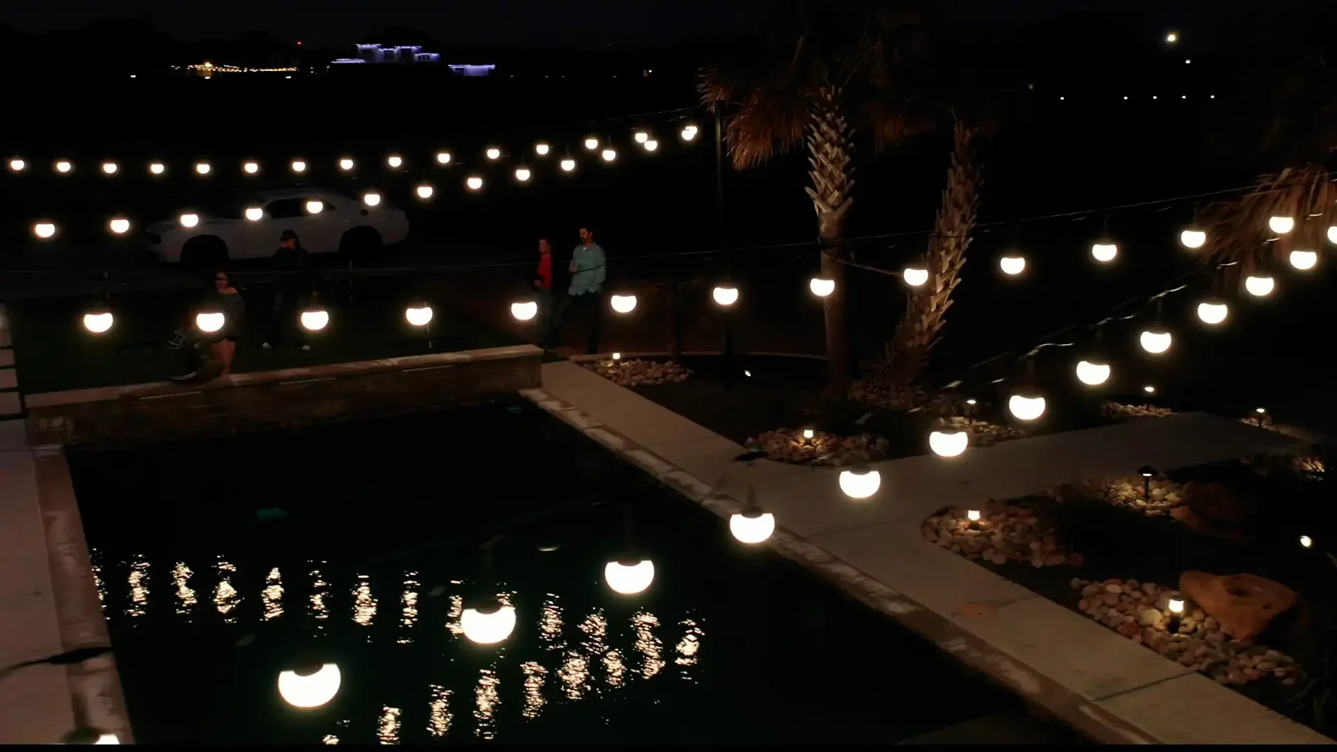 Nighttime scene with hanging patio lights outlined along pathways and trees, with a small water feature reflecting the lights, and a few people walking nearby.