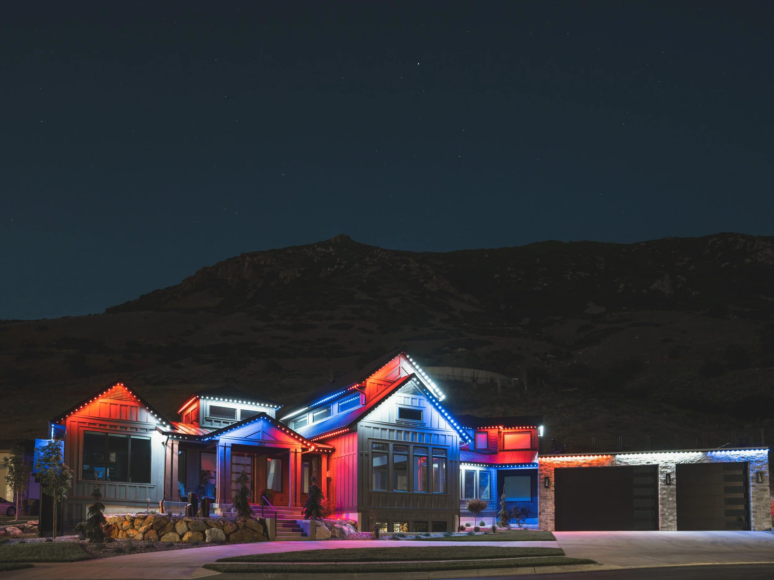 A house illuminated with red, white, and blue eave lights at night, with mountains and a starry sky in the background.