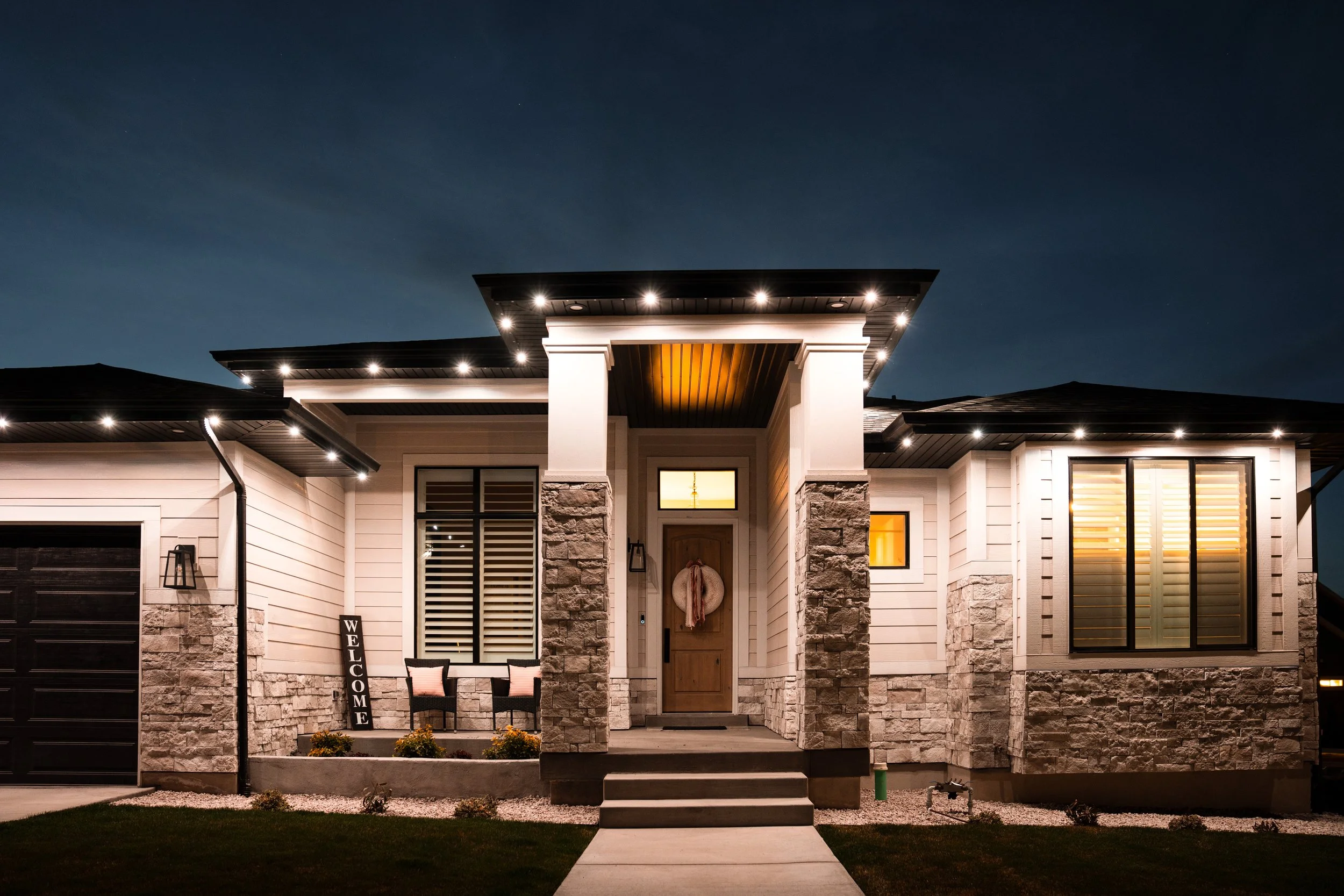 Modern house with stone and siding exterior, illuminated at night, featuring a front porch with steps, chairs, and a welcoming wreath on the door.