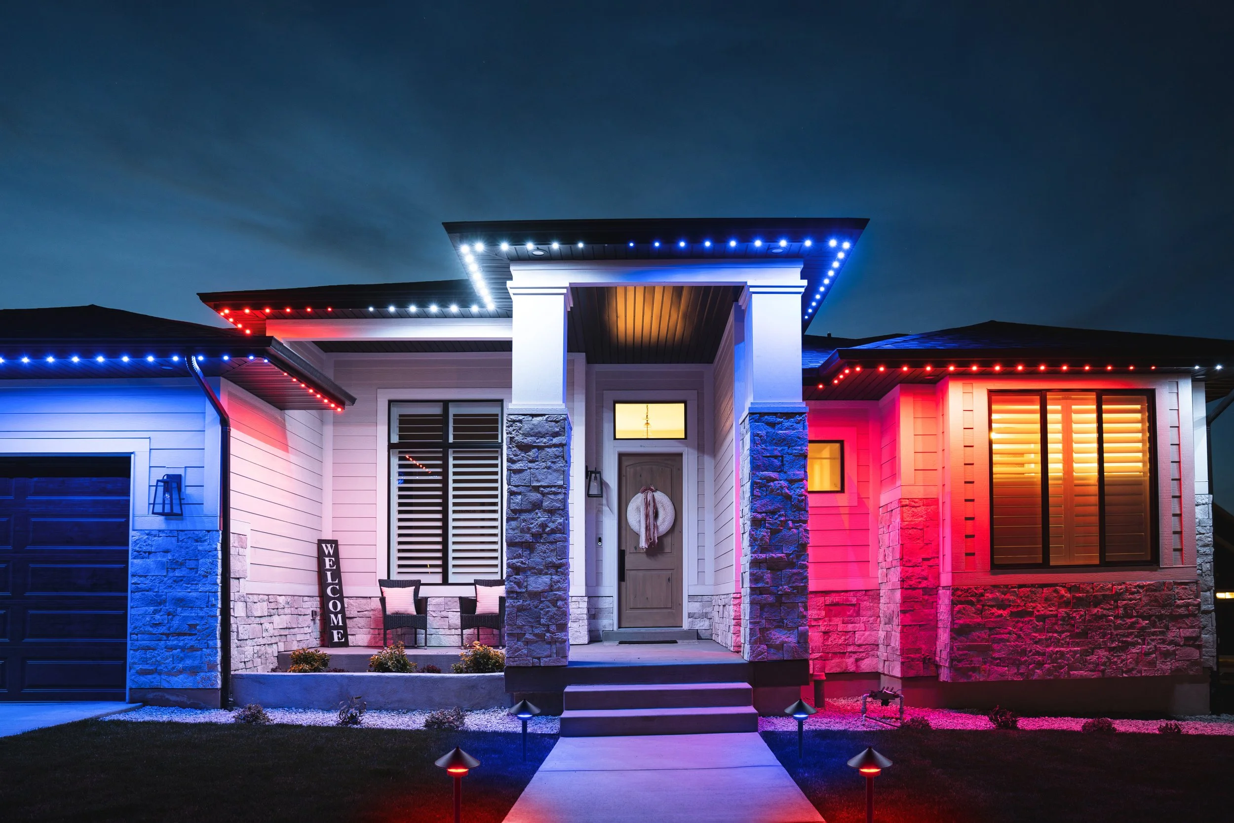 A house decorated with colorful 4th of July lights at night, with a welcome sign and two chairs on the front porch.