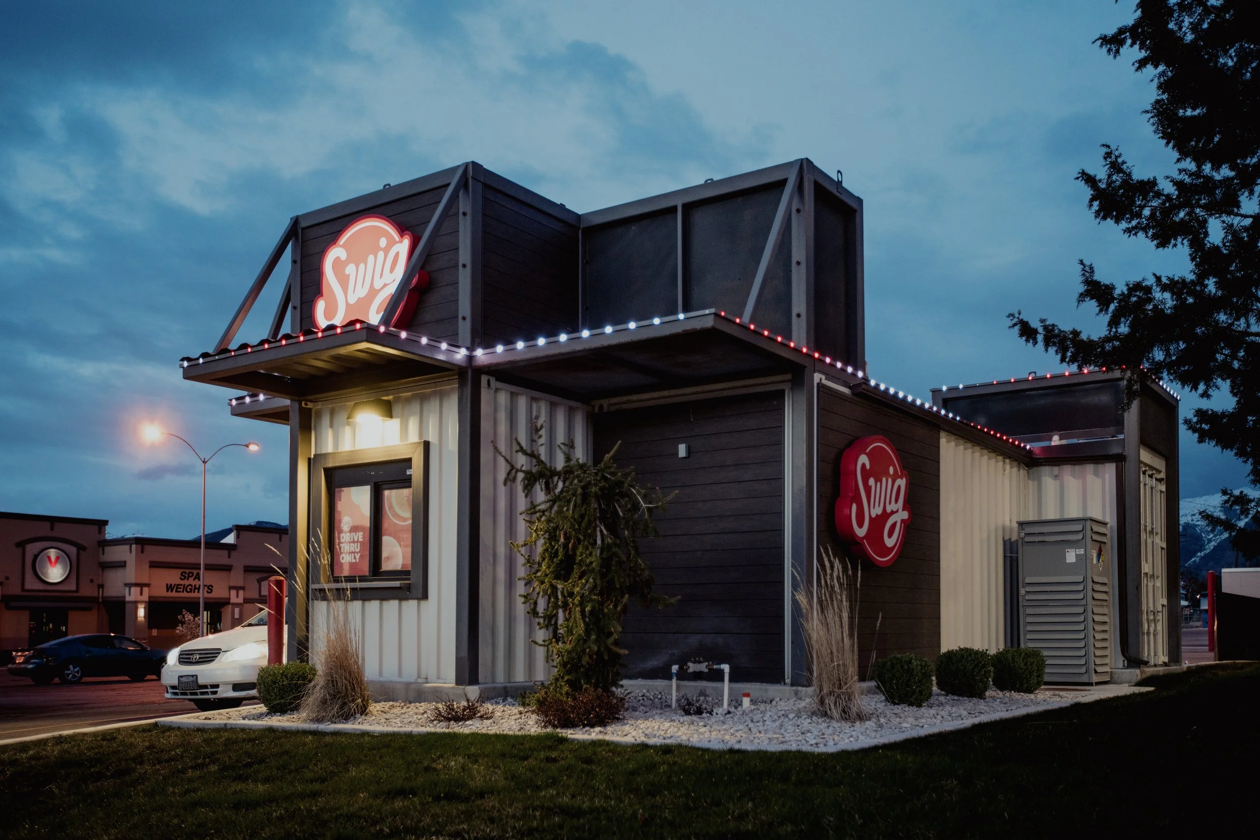 A modern soda shop with black and white siding, illuminated signs, JellyFish Lights, and a drive-thru window at dusk.