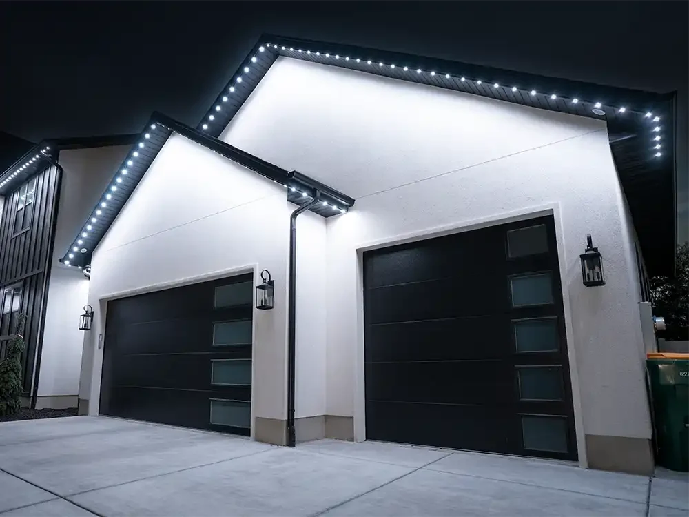 Modern duplex garage with black doors, white exterior walls, and security eave lighting at night.