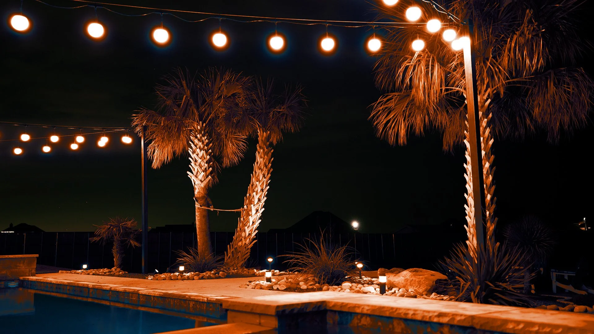 Nighttime scene of a backyard with patio lights hanging overhead, tall palm trees illuminated with orange lighting, small landscape lights, and a swimming pool.
