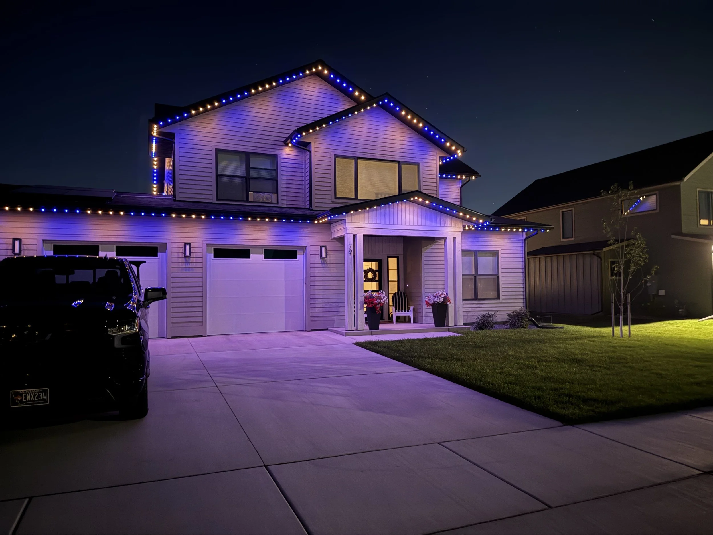 A two-story house decorated with colorful blue and orange lights at night. The house has a driveway with a black vehicle parked on it, front porch with two benches and potted flowers. The yard is well-maintained with grass and small trees.