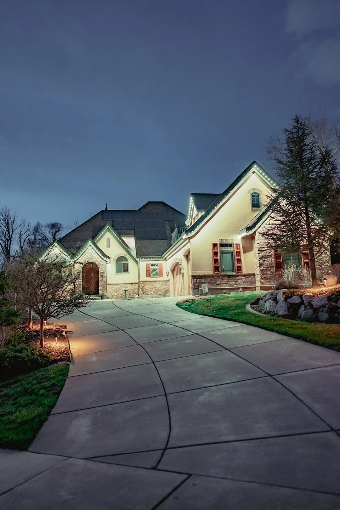 A large house illuminated with JellyFish Lighting at night, featuring a curved driveway, landscaped yard with rocks and trees, and a dark sky above.