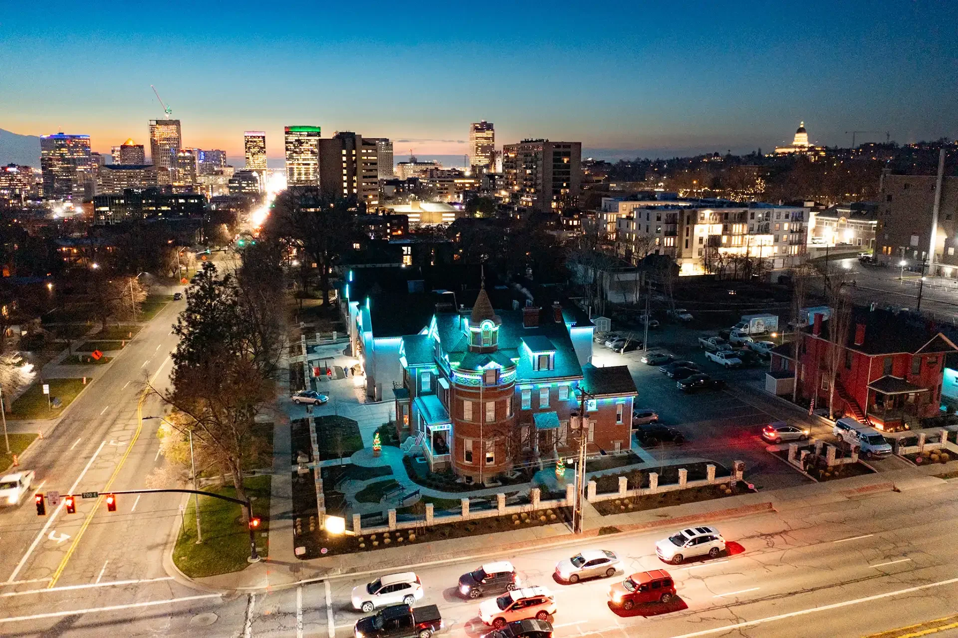A cityscape at dusk showing a historic house with colorful neon lighting in the foreground, with modern high-rise buildings and the Capitol dome in the distance.