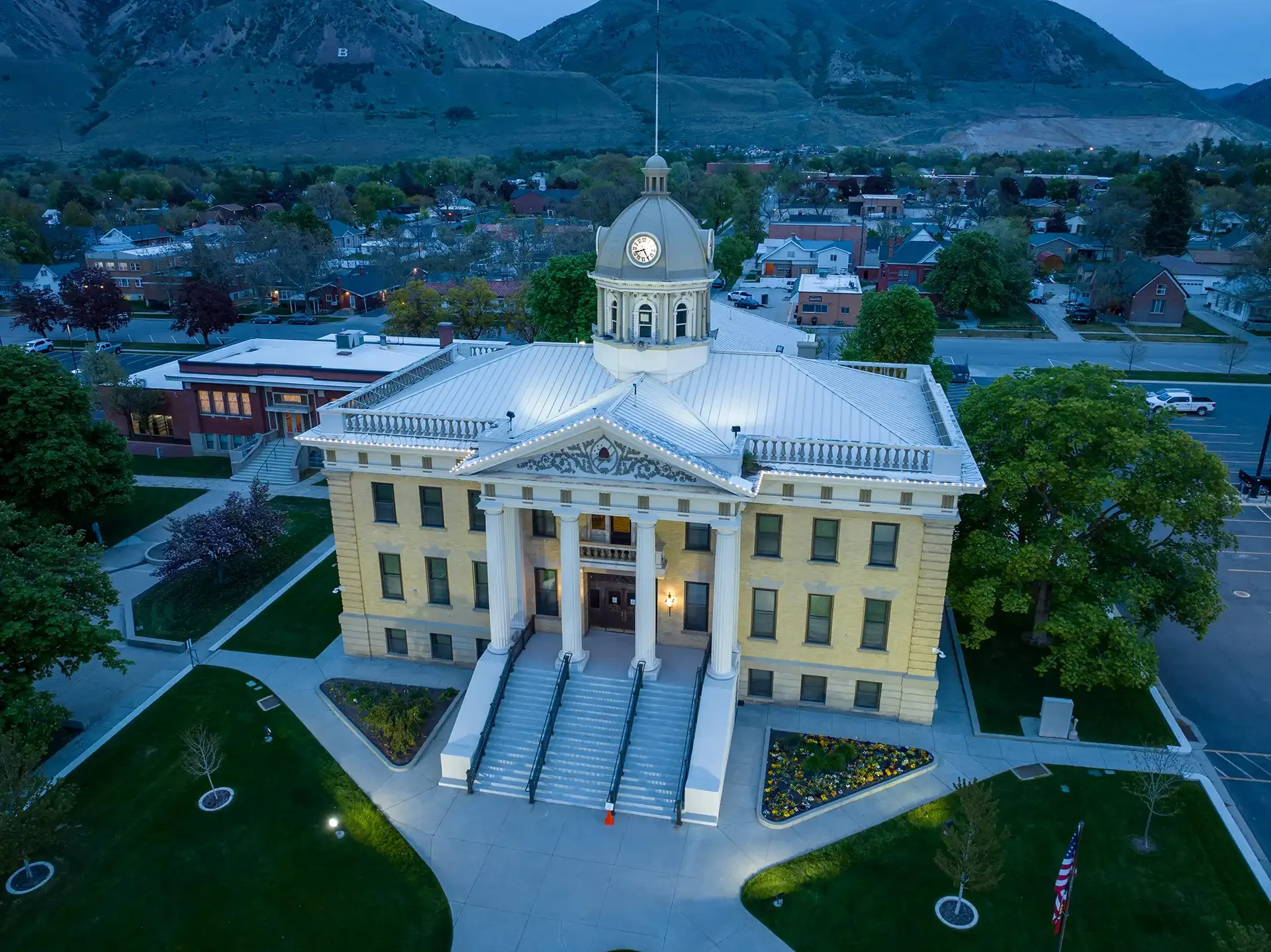 A historic government building with a prominent clock tower, large white columns at the entrance, and a staircase leading up to the front door, surrounded by well-maintained lawns and landscaping, with residential houses and mountains in the background.