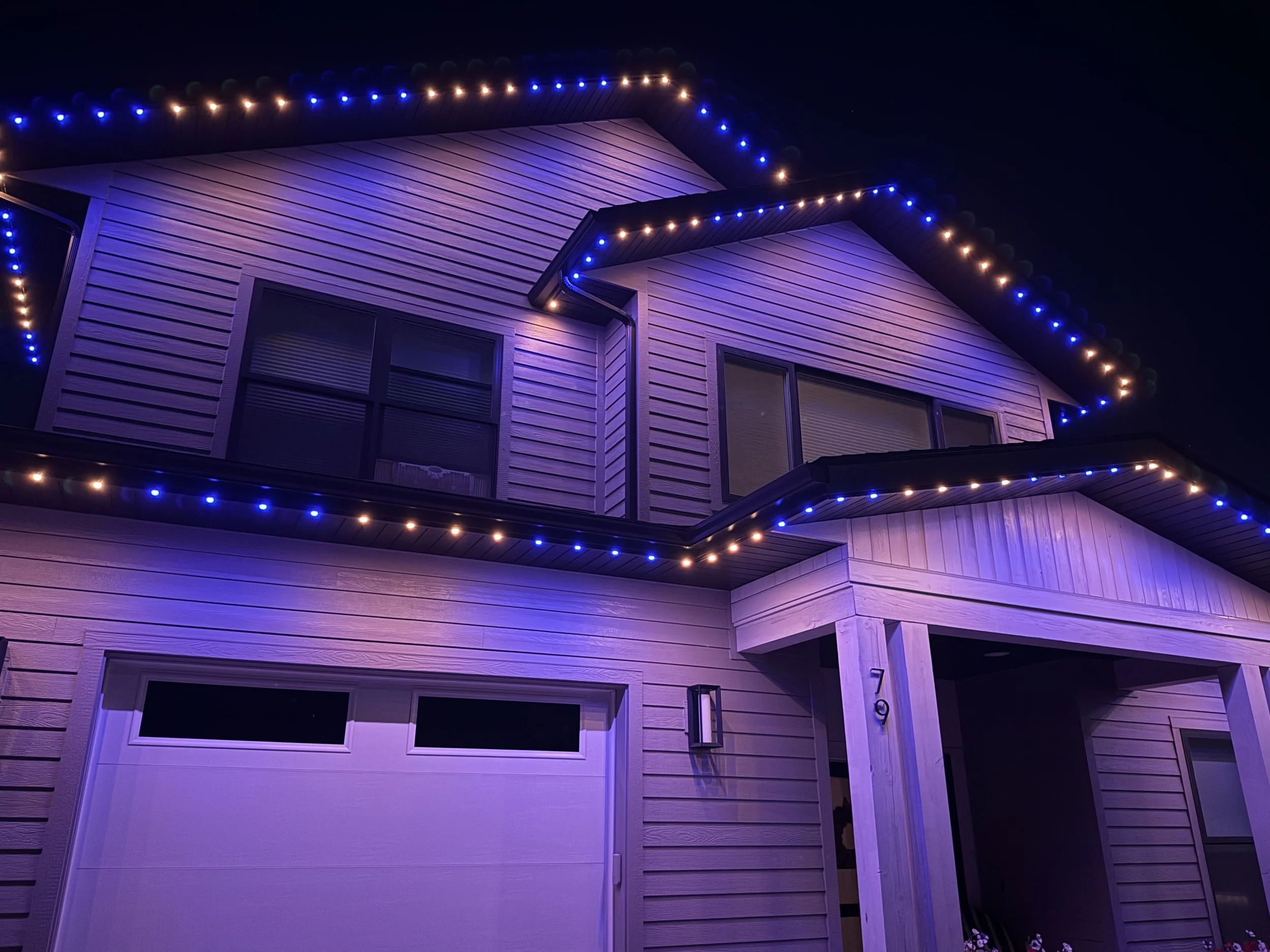 A two-story house decorated with blue and orange eave lights at night, featuring a garage door and front porch.