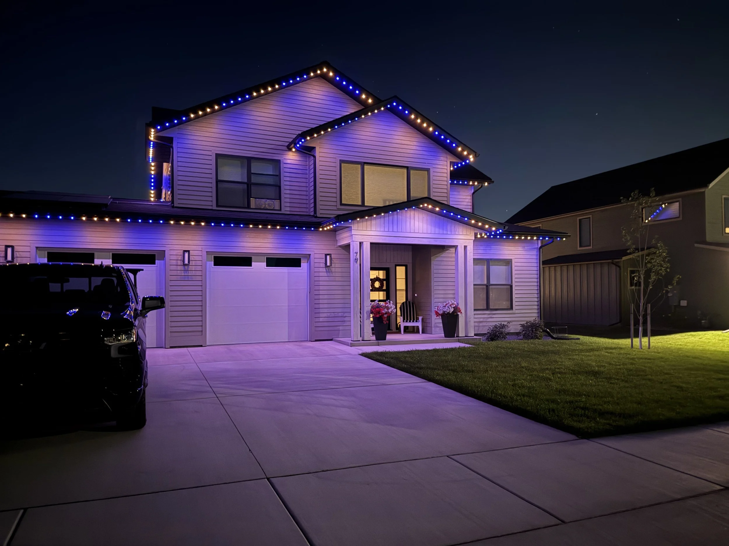 A two-story house decorated with blue and orange JellyFish Lighting at night, with a driveway, a black car parked, and a small front lawn with a tree and potted flowers.