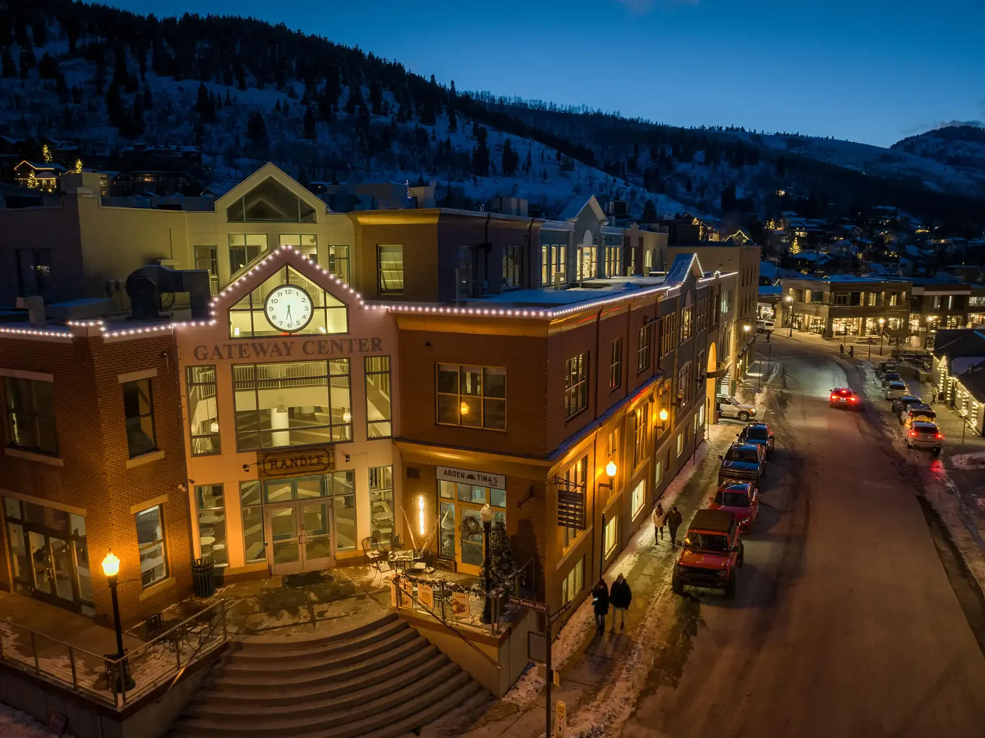 Nighttime view of a small downtown area with illuminated buildings, parked cars, and snow on the ground, with a mountain in the background.