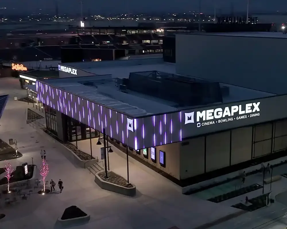 Aerial view of the MegaPlex entertainment center at night with bright purple vertical  JellyFish Saber lights on the exterior walls and illuminated sign displaying movies, bowling, games, and dining.
