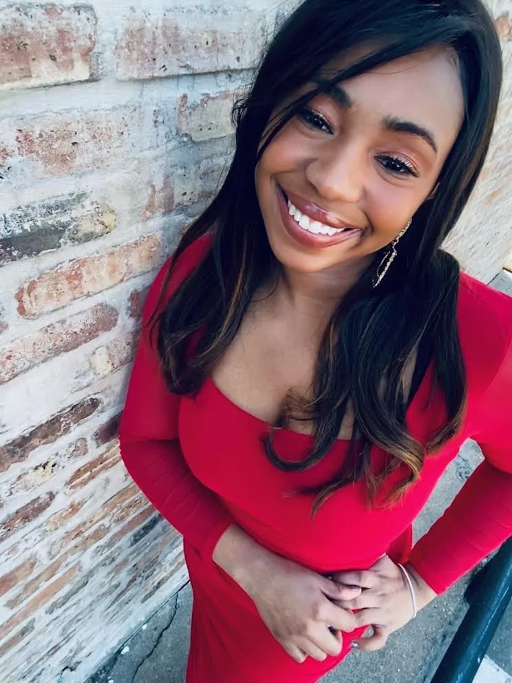 A young woman with long dark hair, wearing a red dress, smiling and standing against a brick wall.