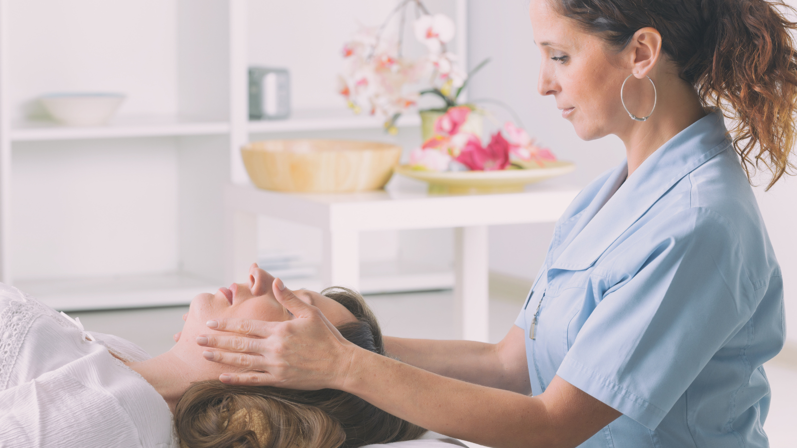 A woman receiving facial treatment from a practitioner in a spa or wellness center with flowers and bowls in the background.