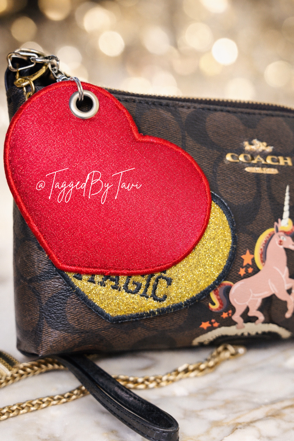 A brown Coach handbag with a pattern and the word 'COACH' on it. A red, heart-shaped patch with text '@TaggedByTavi' stuck to the bag. The patch has a yellow glittery base with the word 'MAGIC' and a pink unicorn sticker behind it. A gold chain and part of a zipper are also visible.