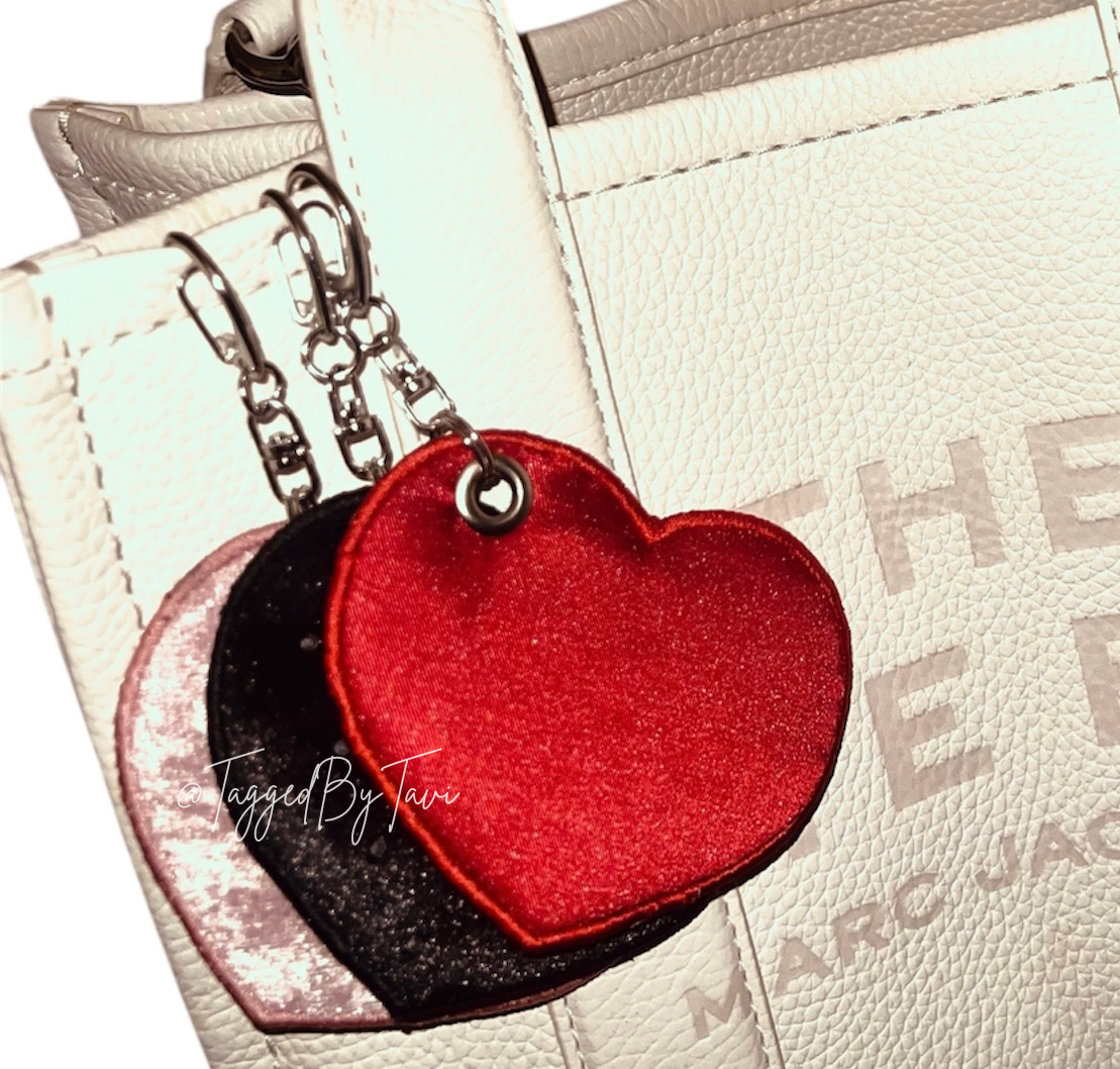 Three heart-shaped keychain accessories in red, black, and pink, hanging from silver chains in front of white textured designer bags.