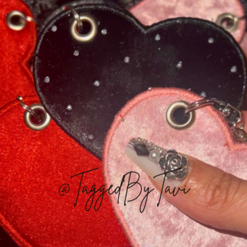 Close-up of two heart-shaped fabric accessories with metal eyelets, one black and the other pink, with a person's finger touching the pink one.
