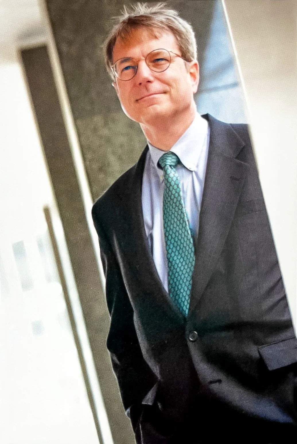 A man in a dark suit, light blue shirt, and patterned teal tie, wearing glasses, stands indoors with a mild smile.