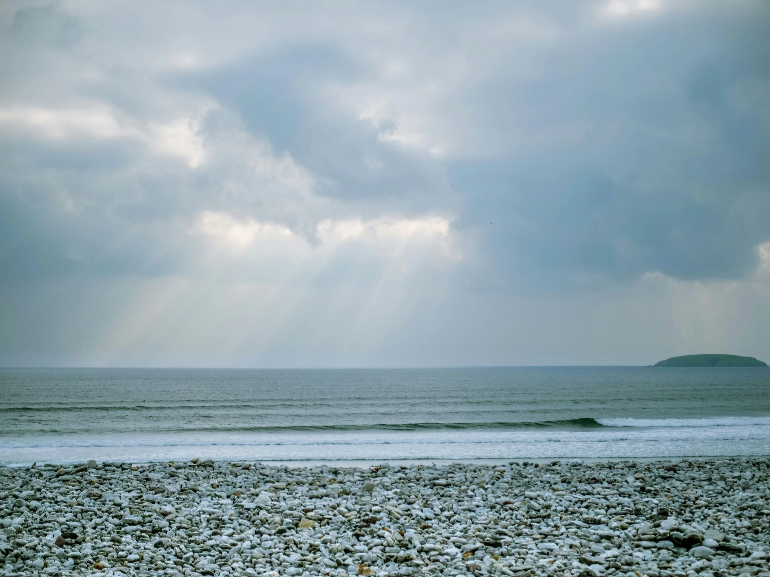 A rocky beach with small pebbles, calm ocean waves, and a cloudy sky with rays of sunlight shining through, in the distance a small island is visible.