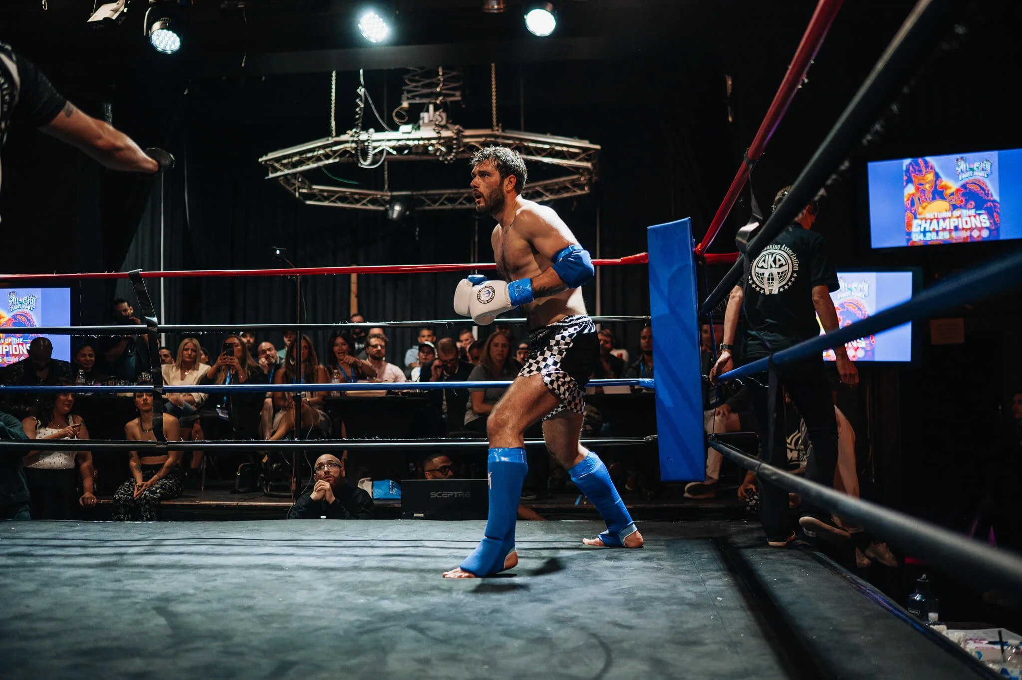 A male Muay Thai fighter in shorts and blue protective gear inside a boxing ring, with an audience watching in the background.