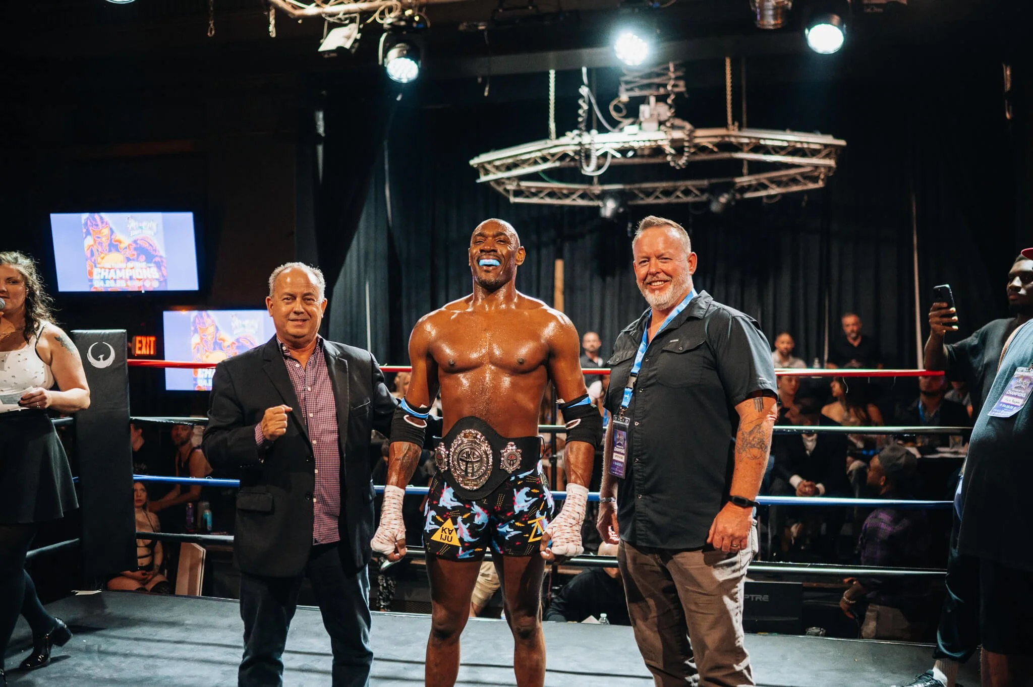 A victorious fighter stands in the ring with a championship belt after winning at All City Fight Night.