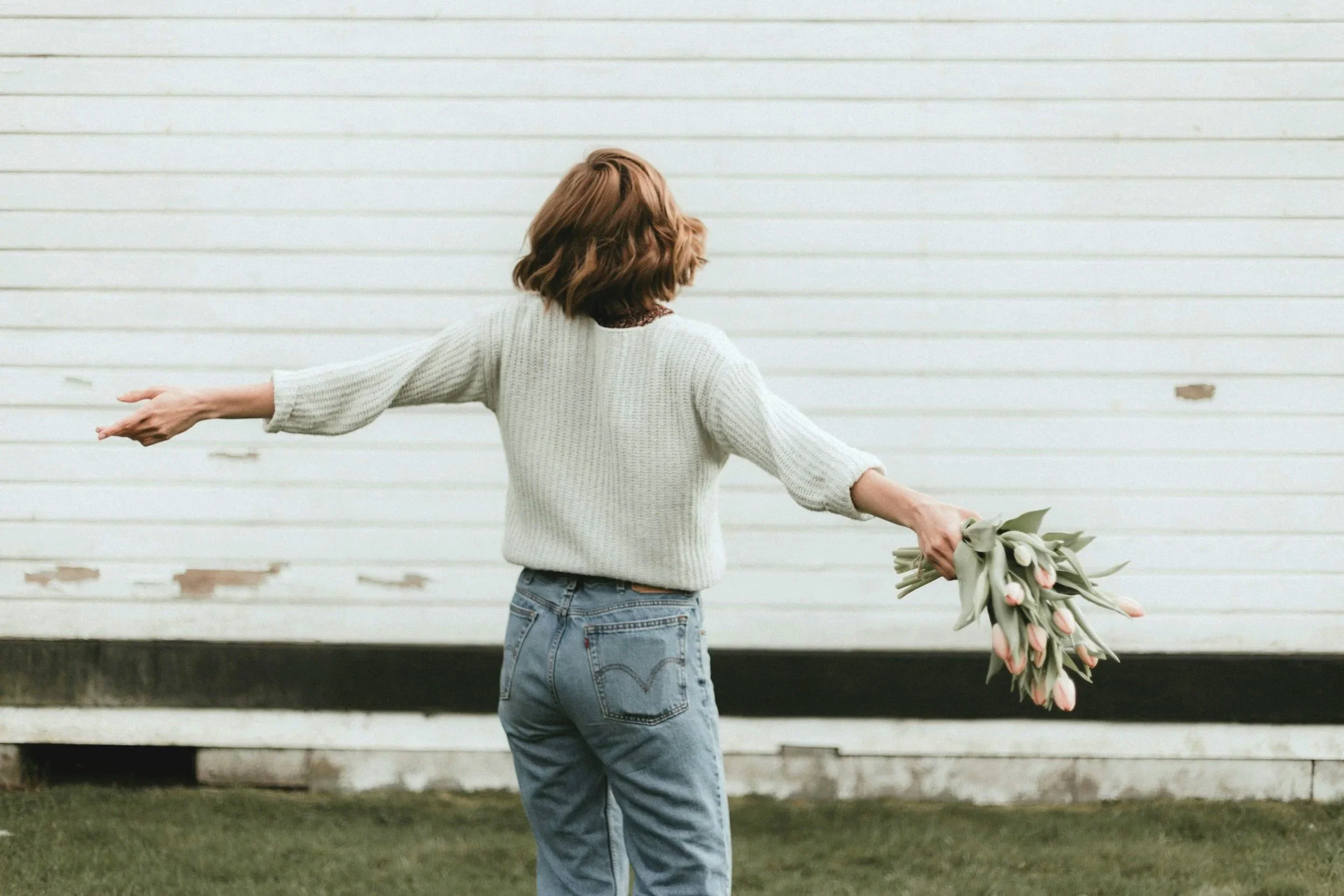 Frau mit braunen Locken in einem weißen Pullover hält einen Blumenstrauß Tulpen in der Hand, während sie vor einer weißen Wand steht.