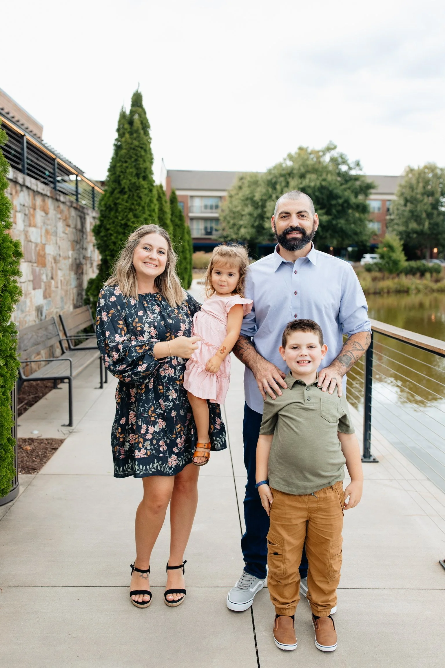 A family of five standing on a sidewalk near a body of water, with trees and modern buildings in the background. The family includes a woman, a man, two boys, and a girl, all smiling at the camera.