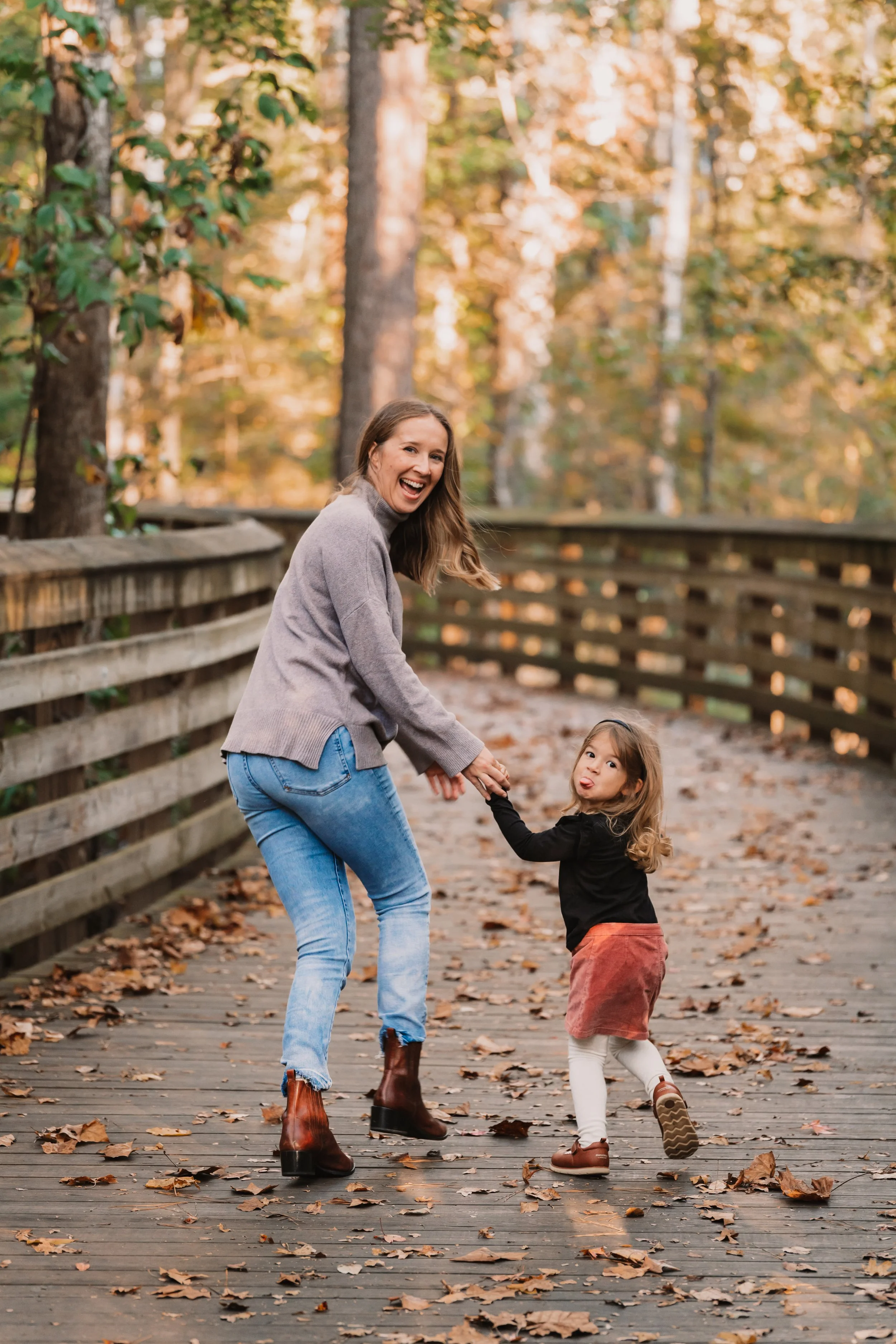 A woman and a young girl are holding hands and playing on a wooden bridge covered in fallen autumn leaves, surrounded by trees with fall foliage.