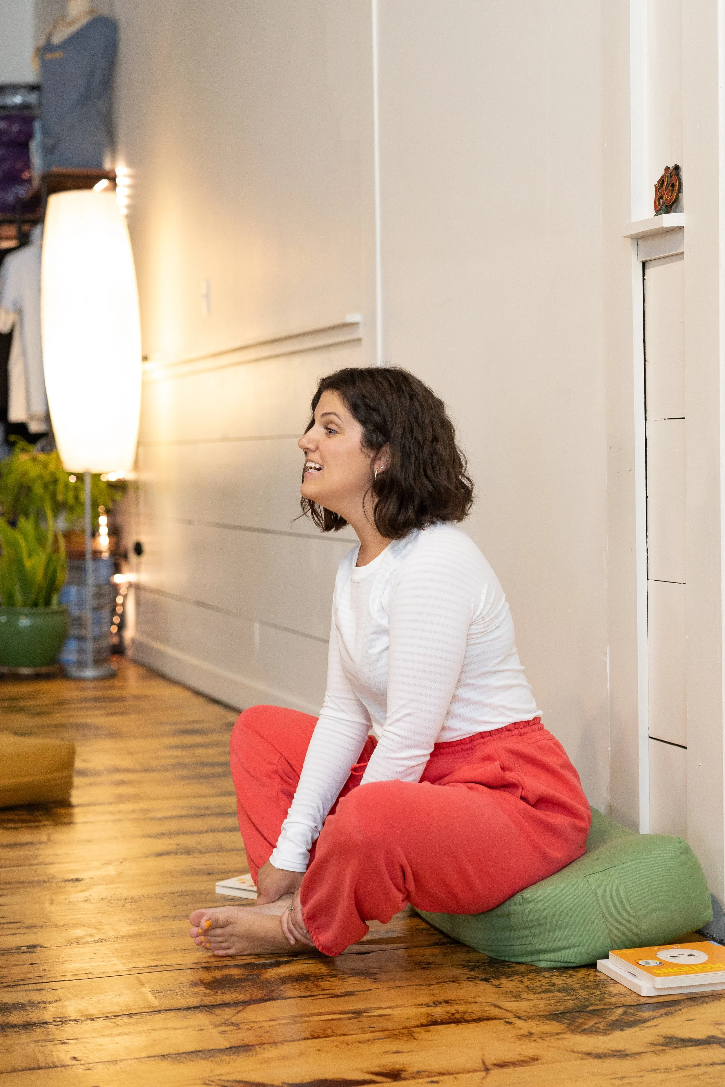 A woman with dark brown, shoulder-length hair, smiling and sitting cross-legged on a green cushion on wooden flooring, wearing a white long-sleeve shirt and red pants, indoors with a white wall and a tall lamp nearby.