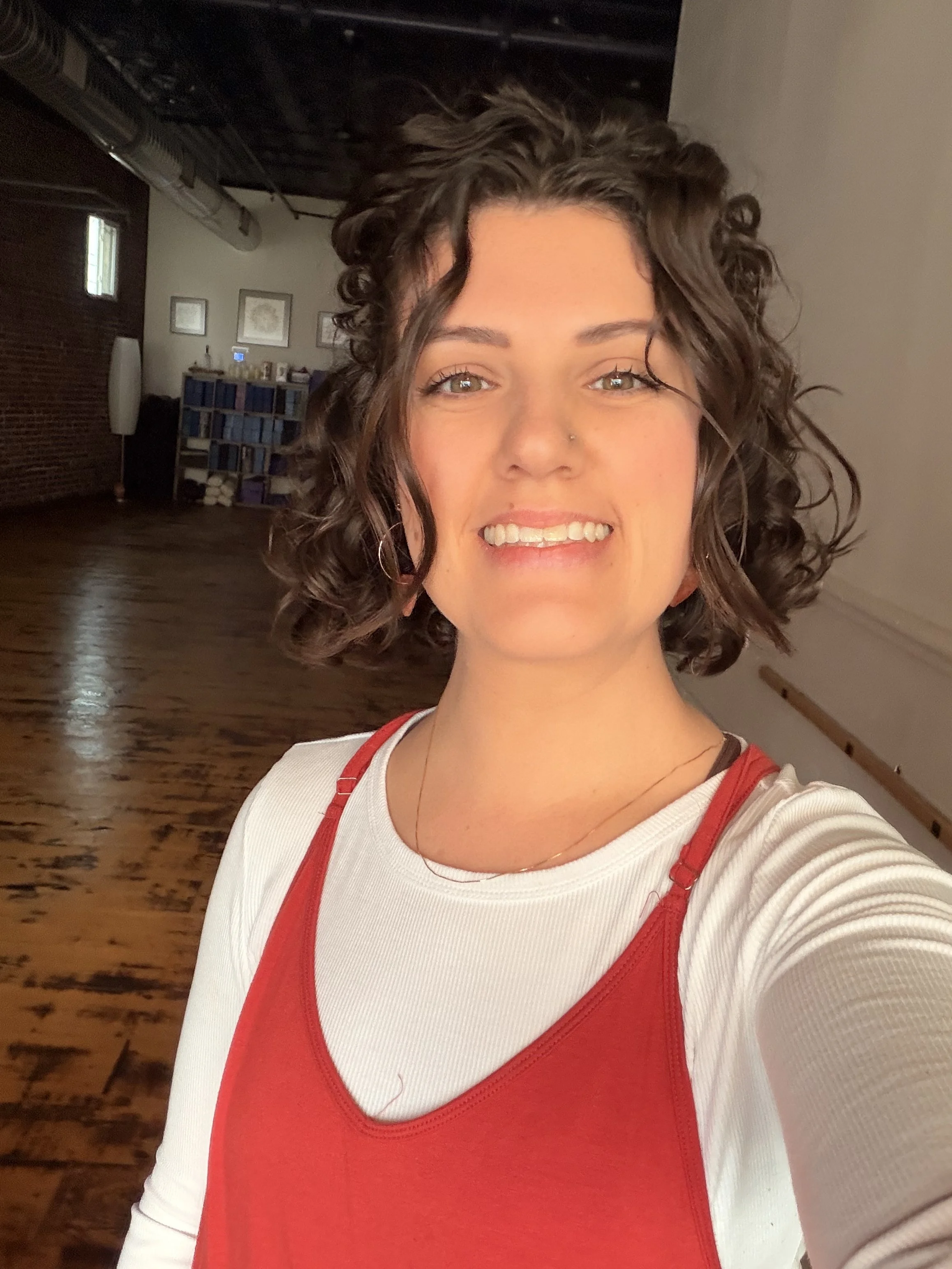 A young woman with curly brown hair taking a selfie in a room with wooden floors and brick walls, smiling at the camera.