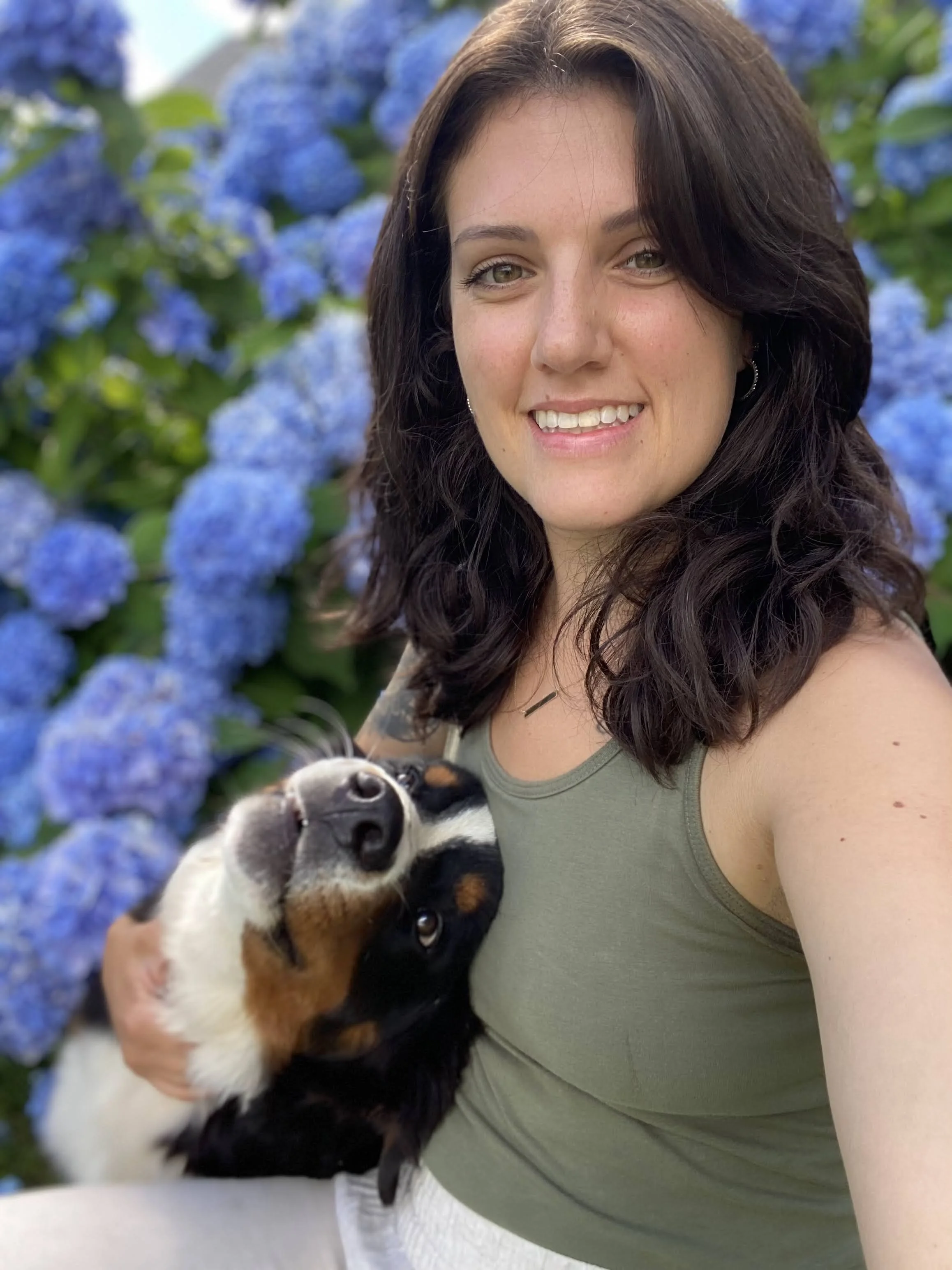 A woman taking a selfie outdoors with blue hydrangea flowers in the background. She is smiling and holding a black, white, and tan dog.